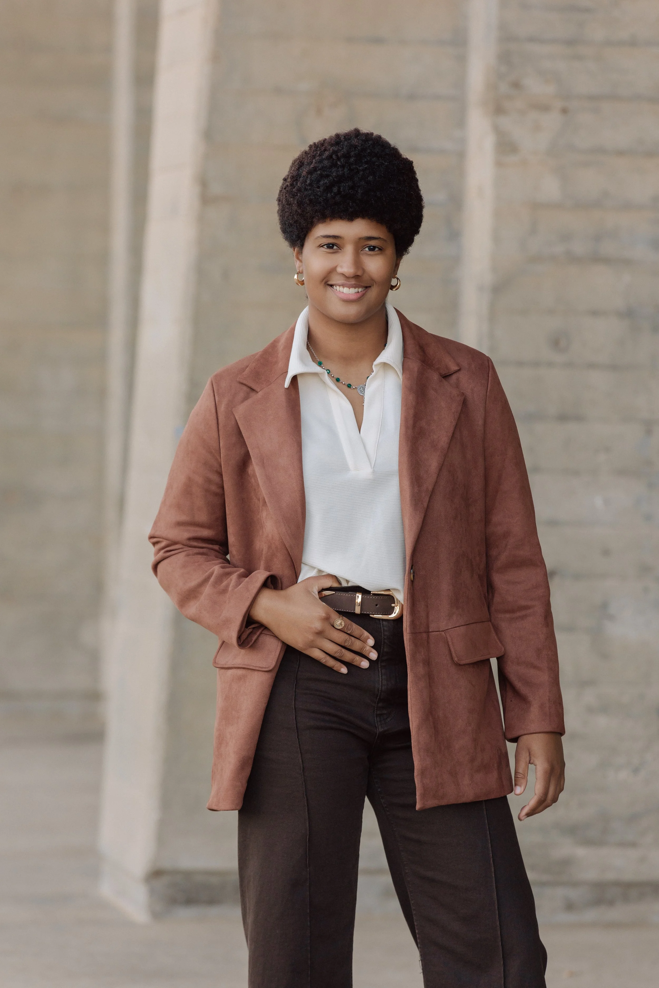 A woman stands outdoors in front of a concrete wall, smiling, wearing a brown blazer, white shirt, and black pants.