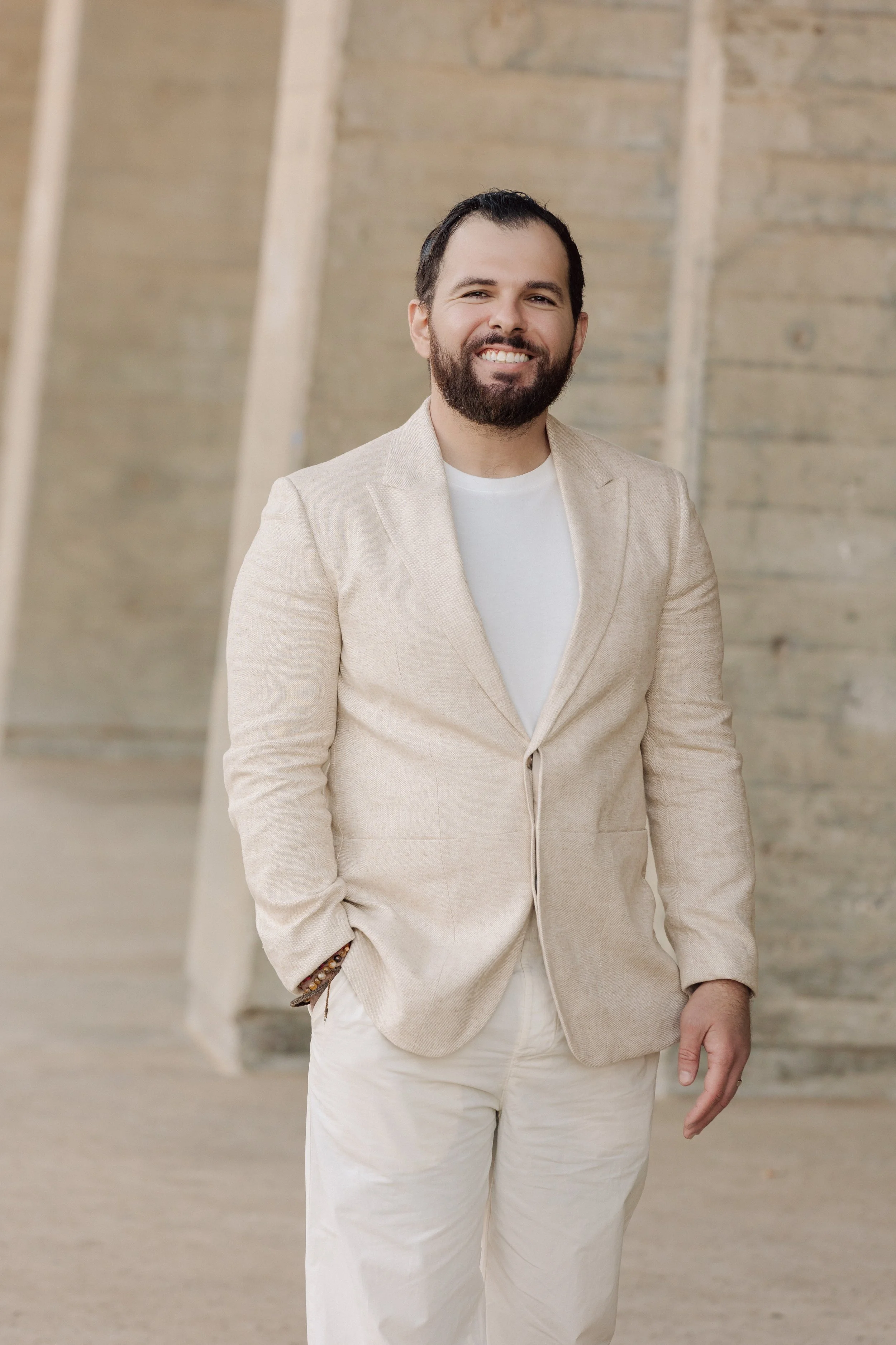 A man with a beard and short dark hair, wearing a beige blazer and white pants, smiling outdoors against a concrete wall.