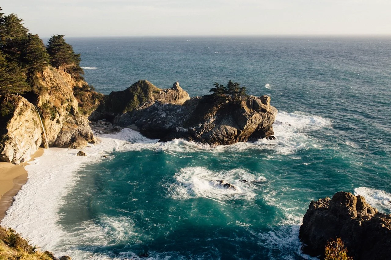 A coastal scene with rocky cliffs, a small beach, and the ocean with waves crashing against the rocks and shoreline.