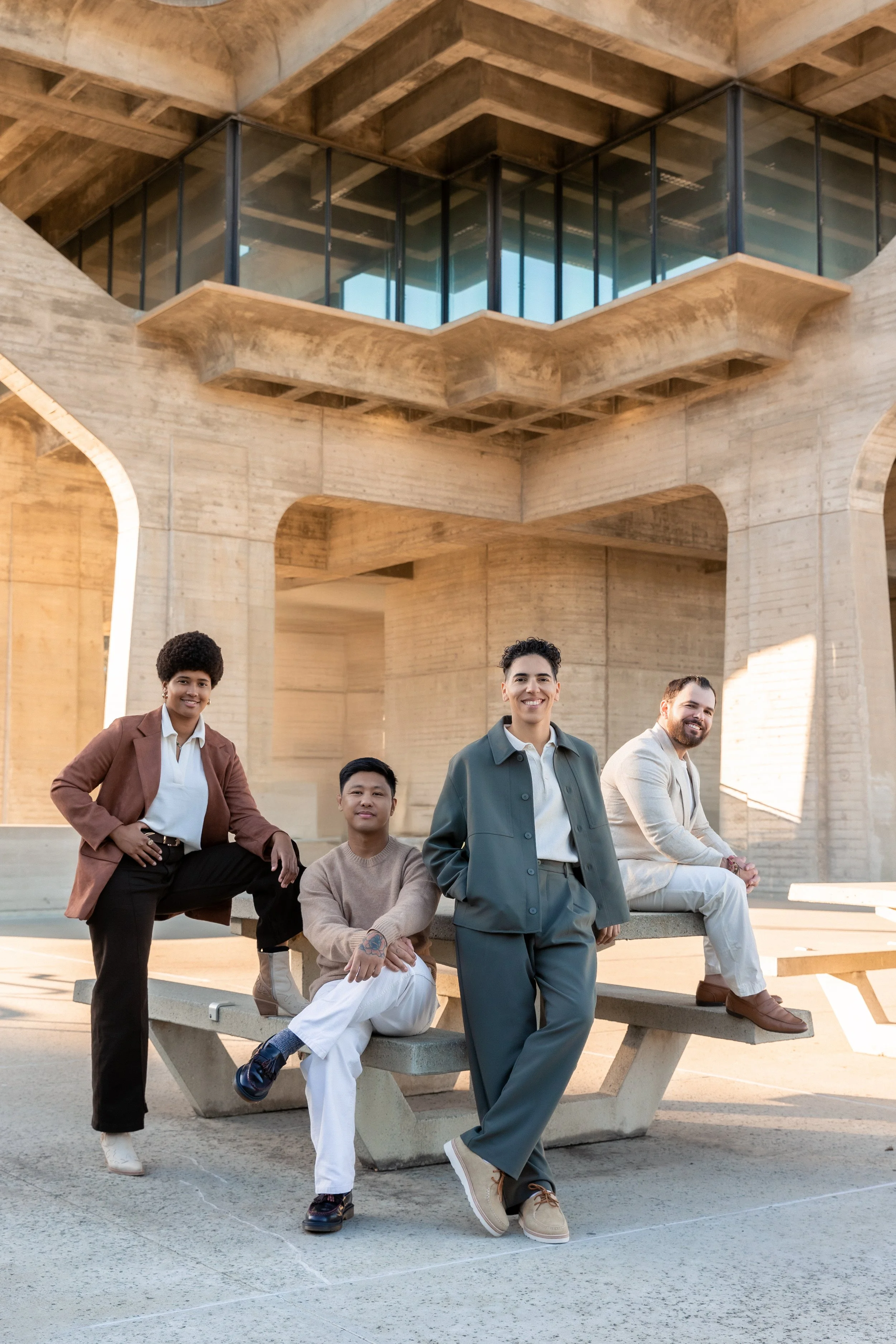A diverse group of four people, two men and two women, gathered outdoors in front of a modern concrete and glass building, smiling and posing for the photo.