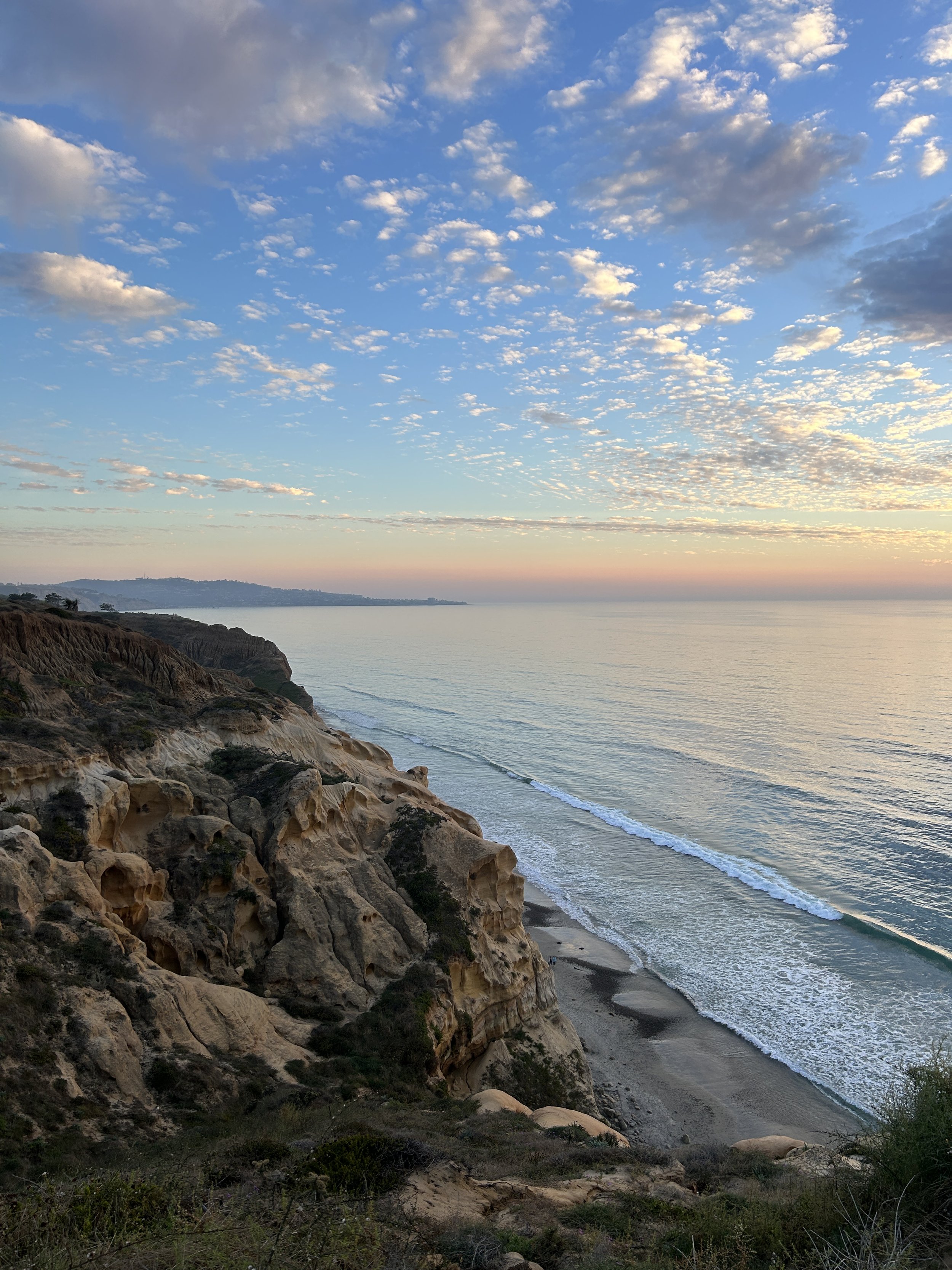 Scenic view of a coastline with cliffs, sandy beach, and ocean under a sky filled with scattered clouds during sunset.