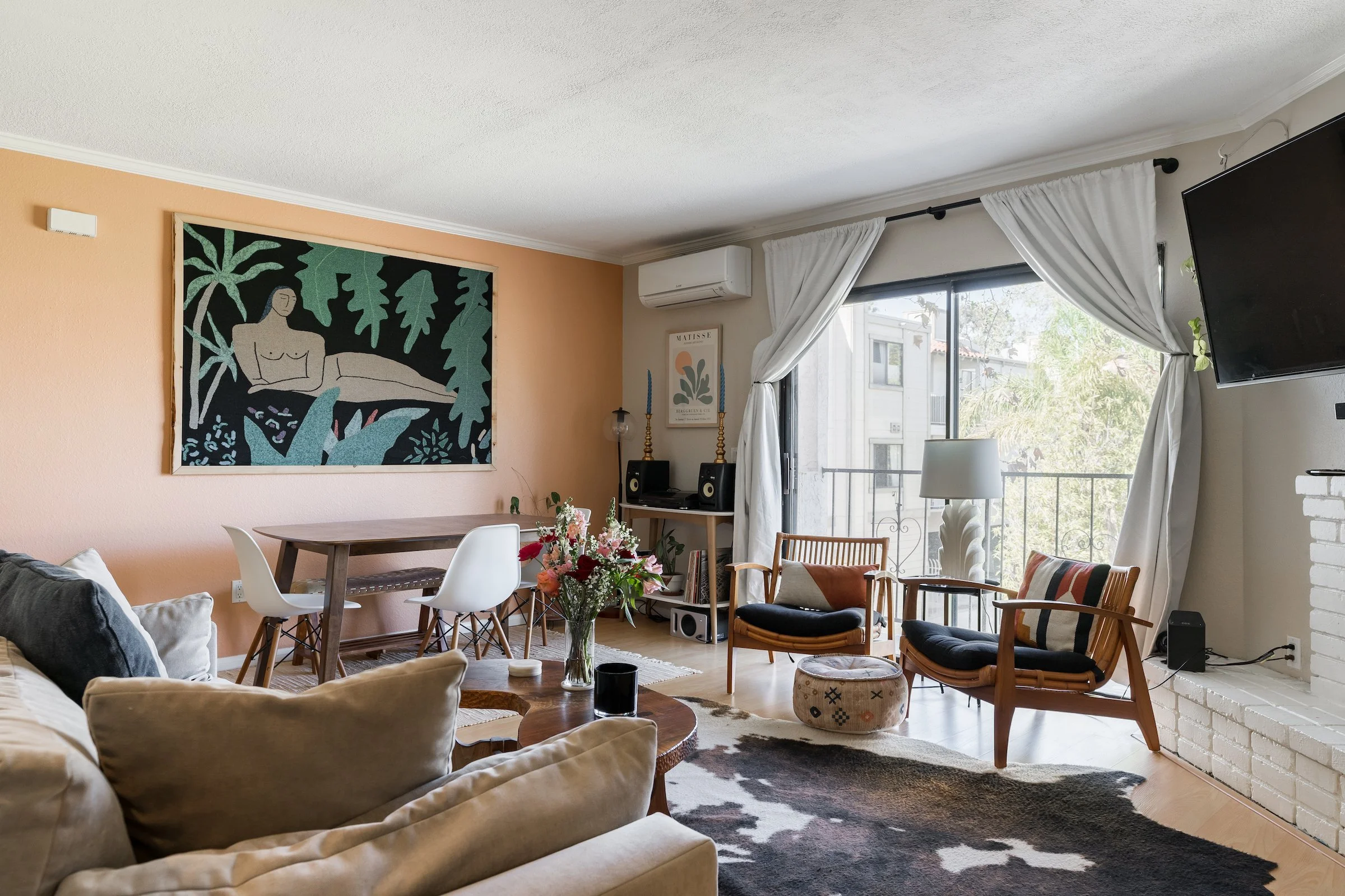 Living room with beige sofa, round wooden coffee table, armchairs near large window with white curtains, wall art, and a television mounted on the wall.