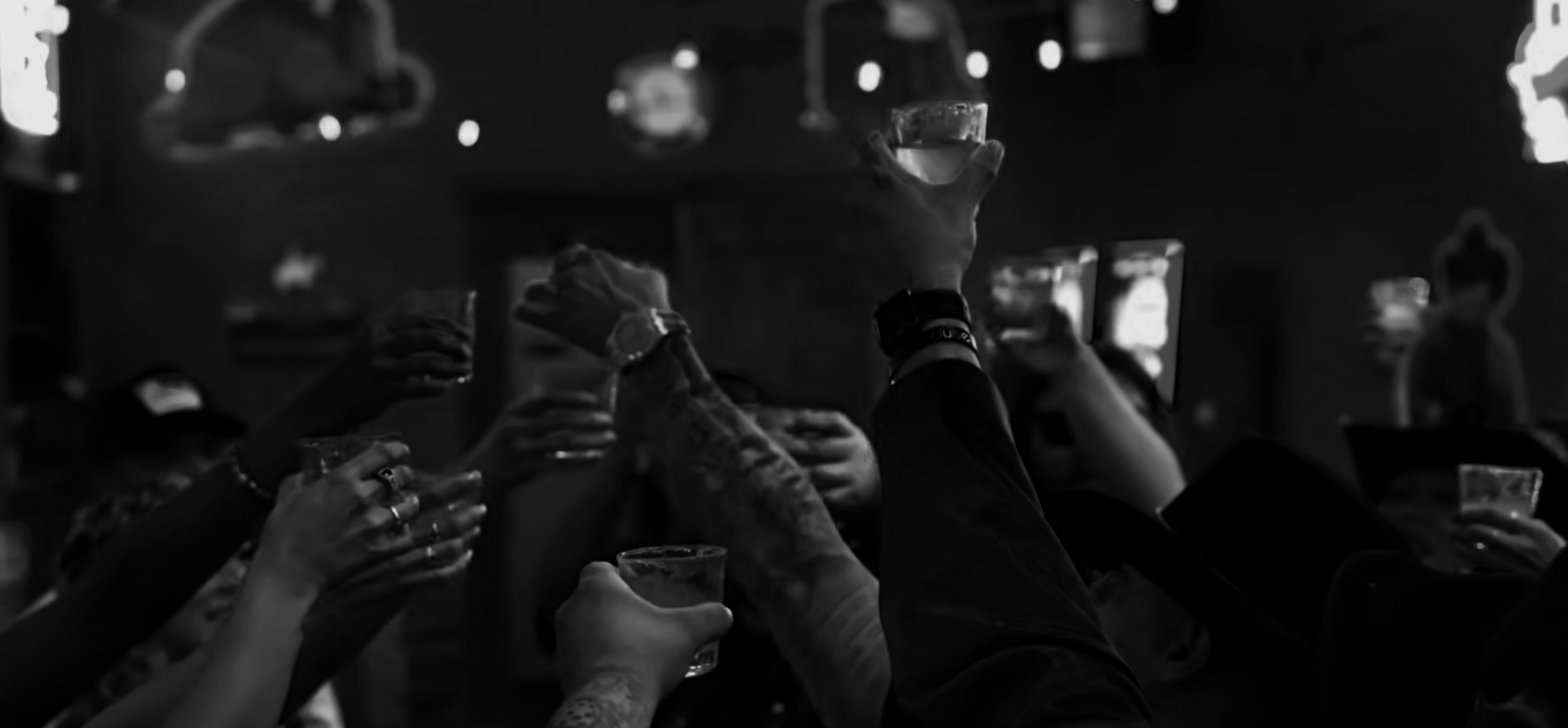 People raise glasses in a toast at a social gathering, with hands and drinks silhouetted against a blurred background.