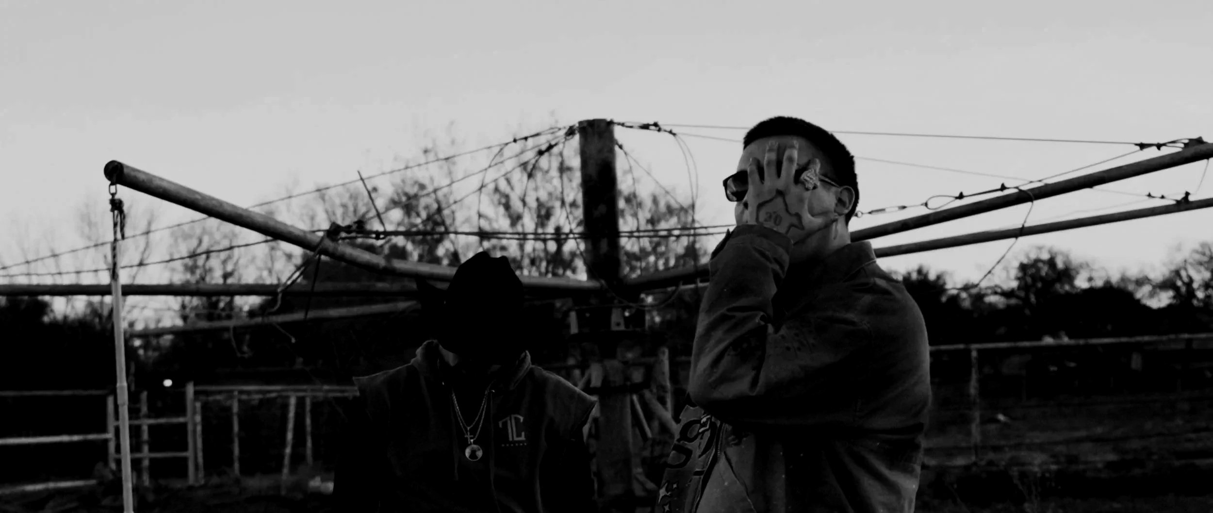 A person with face tattoos wearing sunglasses holding their face with one hand, standing outdoors near scaffolding or construction materials in a black and white photo.