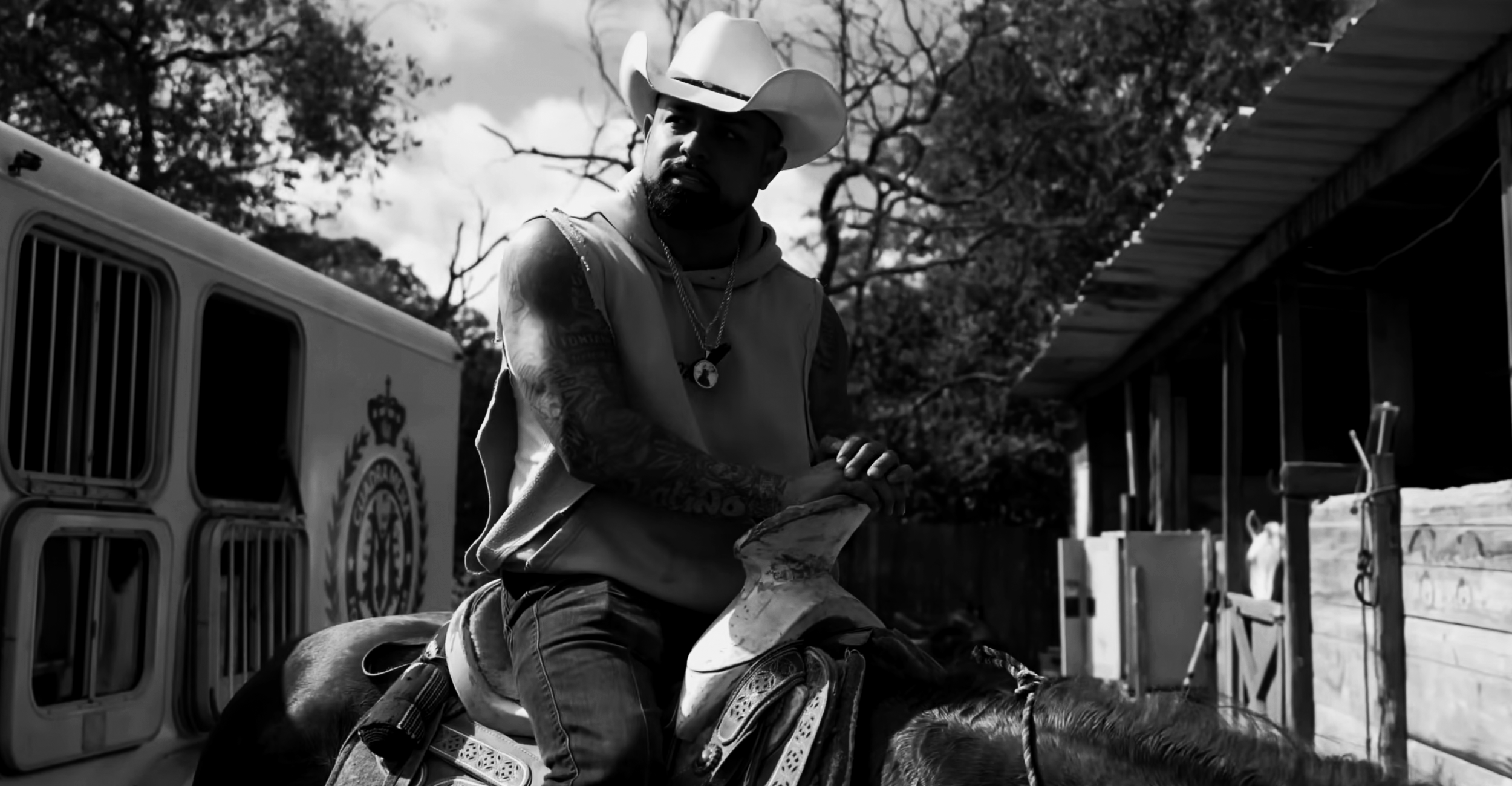 A man wearing a cowboy hat rides a horse, with trees and a building in the background.