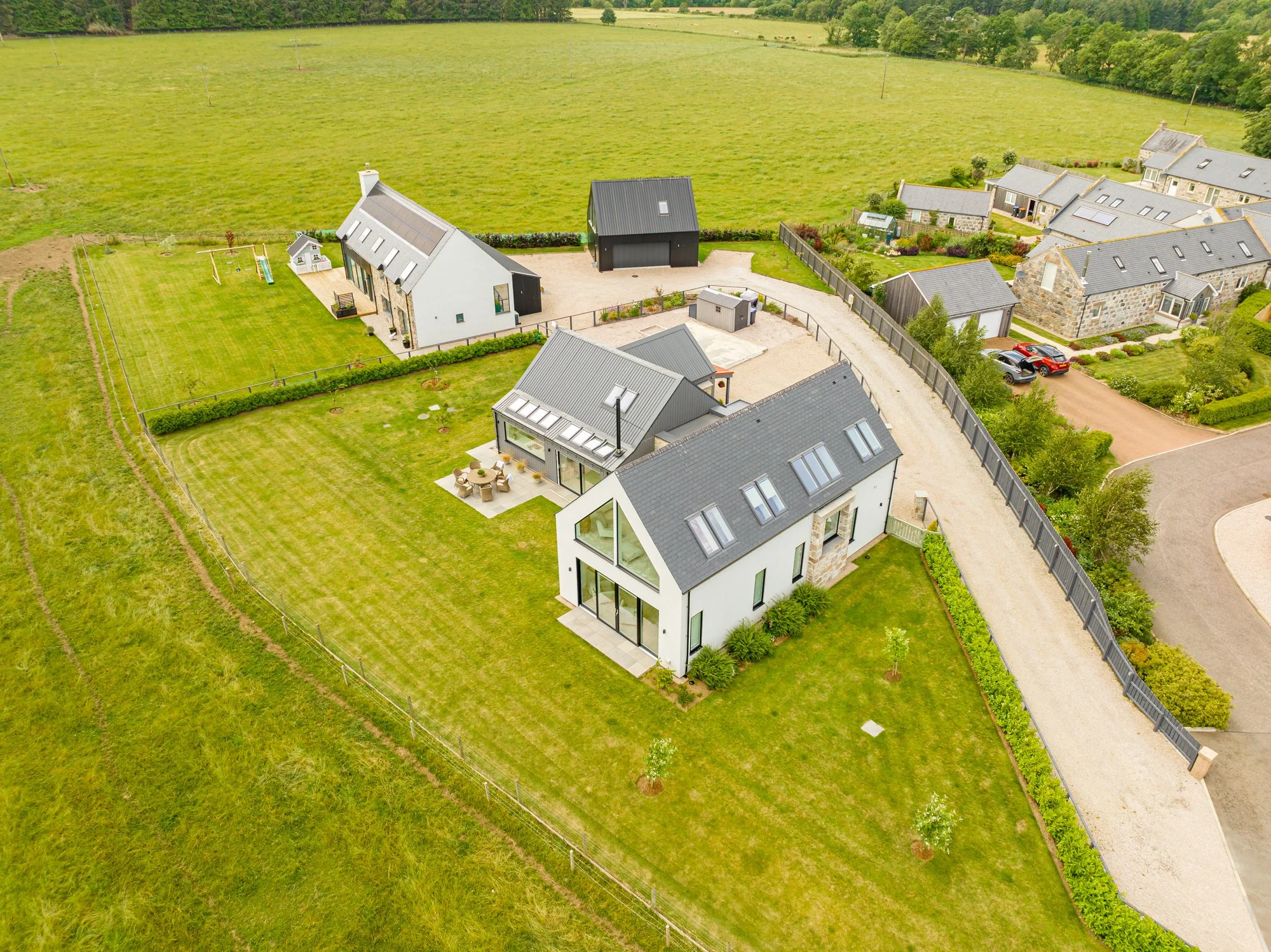 Aerial view of modern houses with gray roofs and a fenced green yard, surrounding a large open green field.