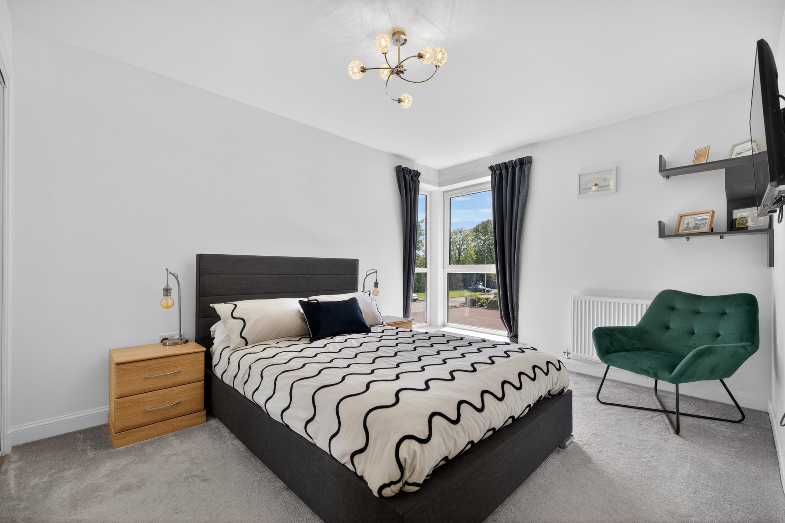 A modern bedroom with a bed, two bedside tables, a teal armchair, large windows with dark curtains, and a ceiling light fixture with multiple globes.