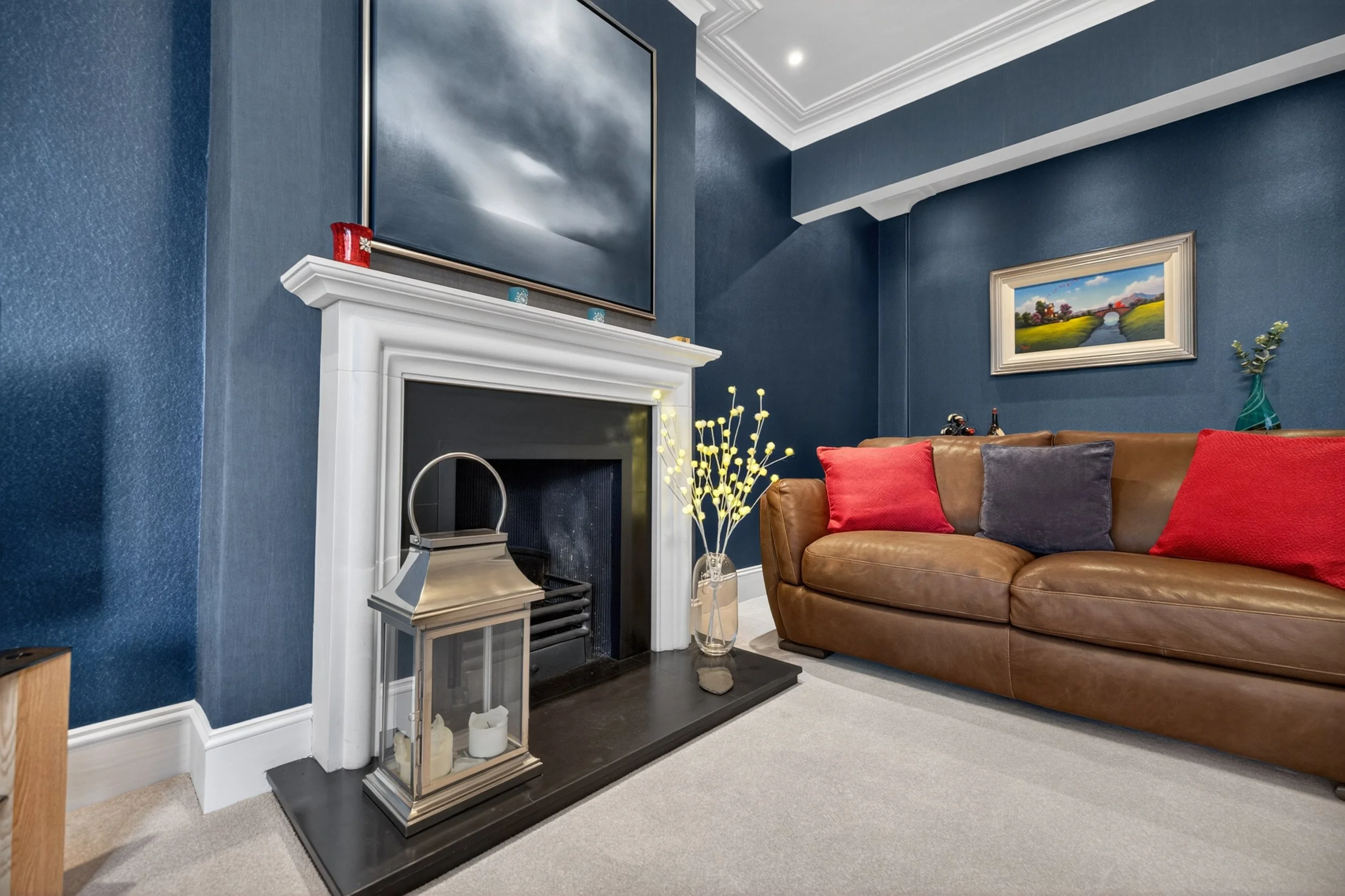 Living room with a brown leather sofa with red and gray pillows, a white fireplace with a black hearth, a glass lantern with candles, a vase with green foliage, a painting of a colorful landscape, and a large TV mounted above the fireplace, with blue