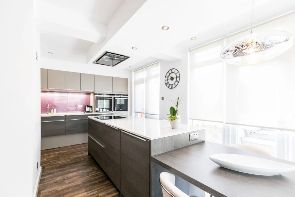 Modern kitchen with light-colored cabinets, a pink backsplash, stainless steel appliances, and a white countertop island. There is a wall clock and large windows with blinds letting in natural light.