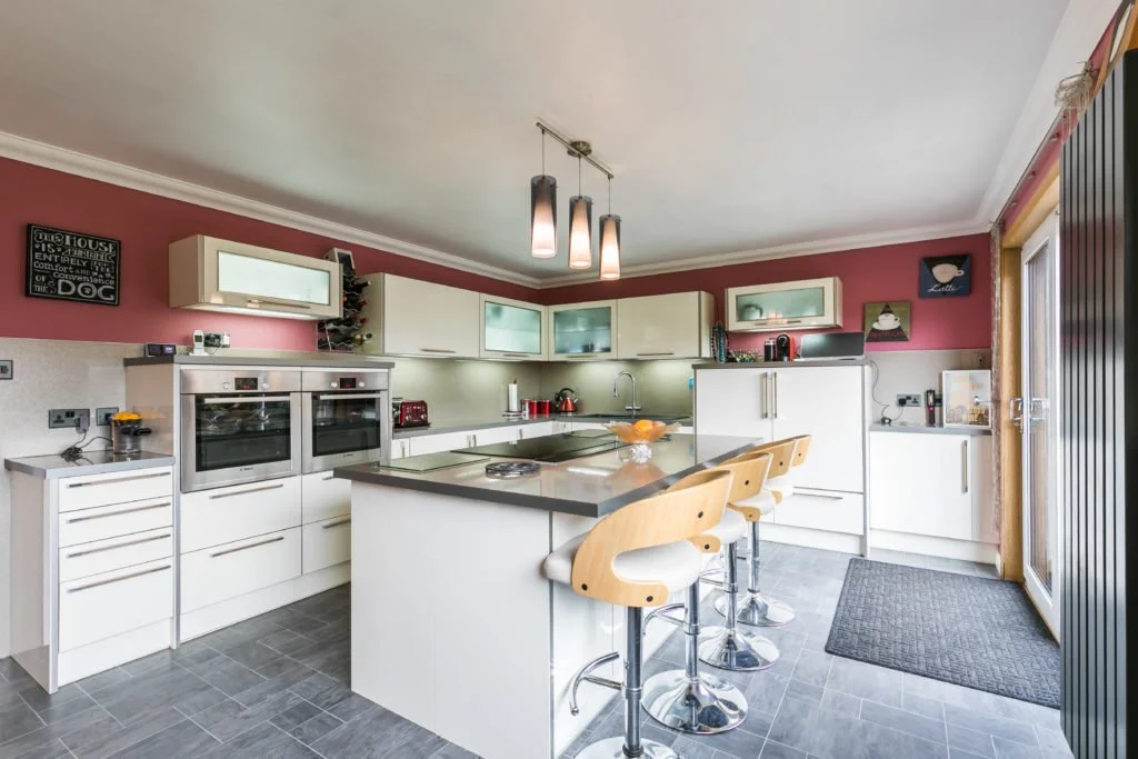 Modern kitchen with white cabinets, a central island with four bar stools, red and beige decor, and stainless steel appliances, illuminated by ceiling lights.