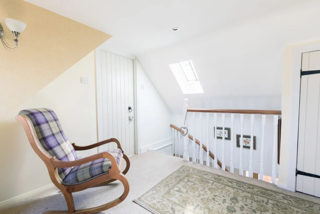 An interior view of a staircase landing with a rocking chair, framed pictures on the wall, a skylight, and a closet door.