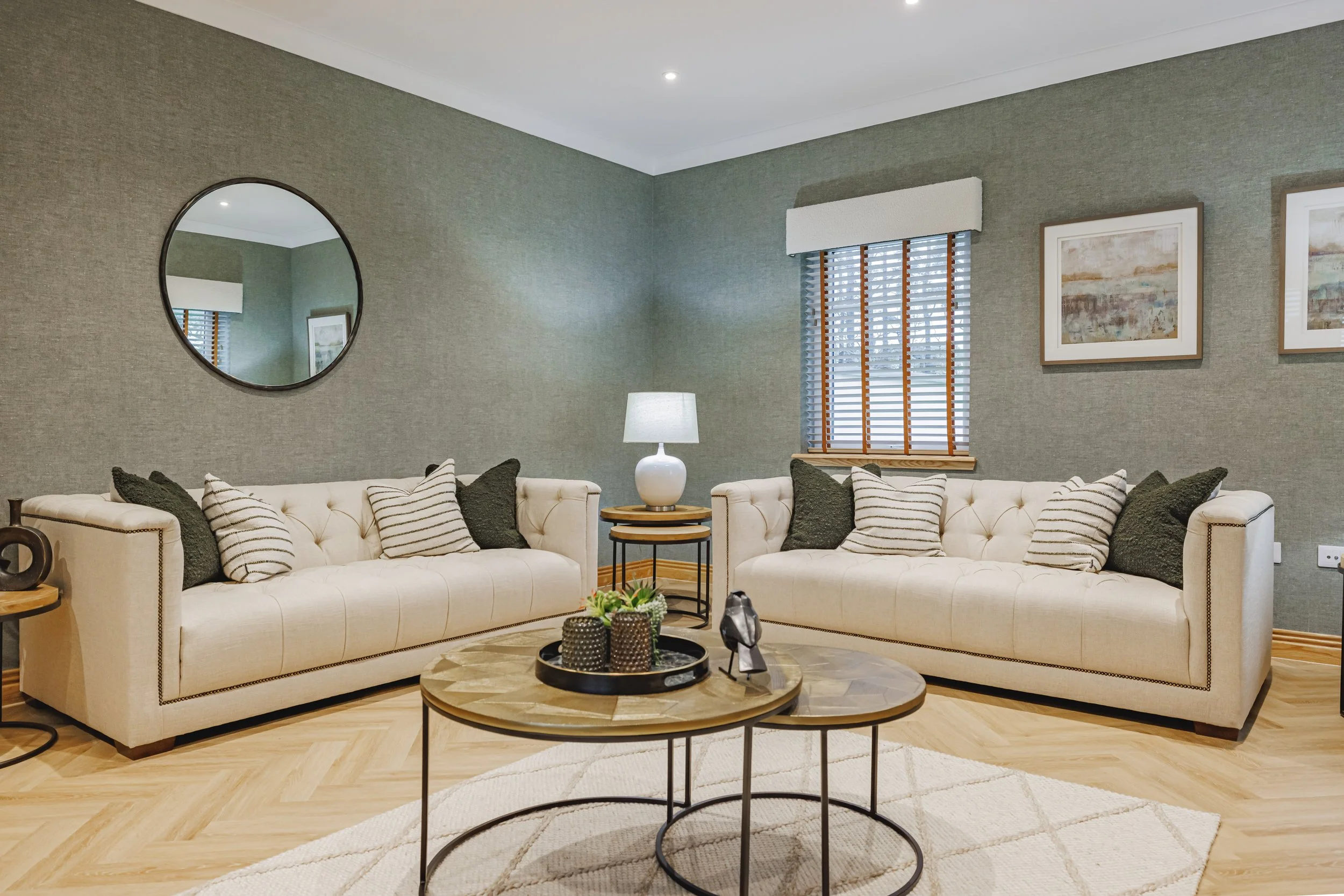 Living room with two cream sofas with striped and dark pillows, round coffee tables, a white lamp, a wall mirror, framed artwork, and a window with blinds.