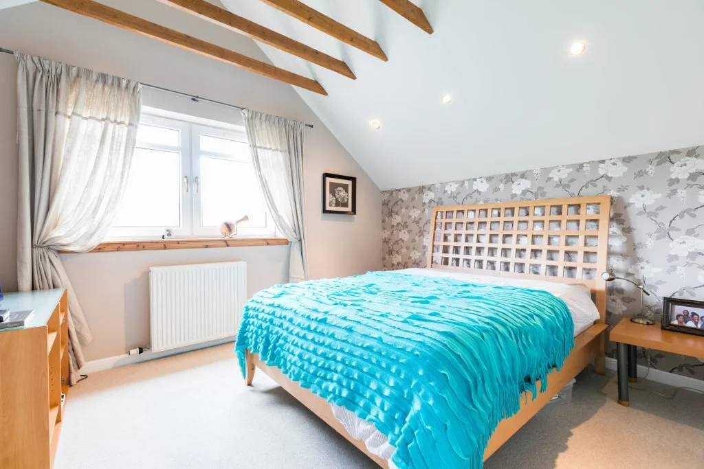 A cozy bedroom with a sloped ceiling, floral wallpaper on one wall, a wooden bed with a blue textured blanket, a bedside table with a lamp and photo frame, a window with cream curtains, and exposed wooden ceiling beams.