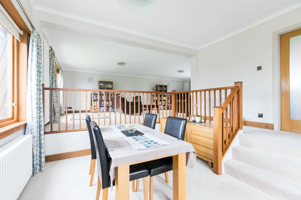 Dining area with four black chairs around a wooden table, a cloth cover, and a table runner, in front of a staircase with a wooden railing, leading to a living room with bookshelves, a hammock, and windows with patterned curtains.