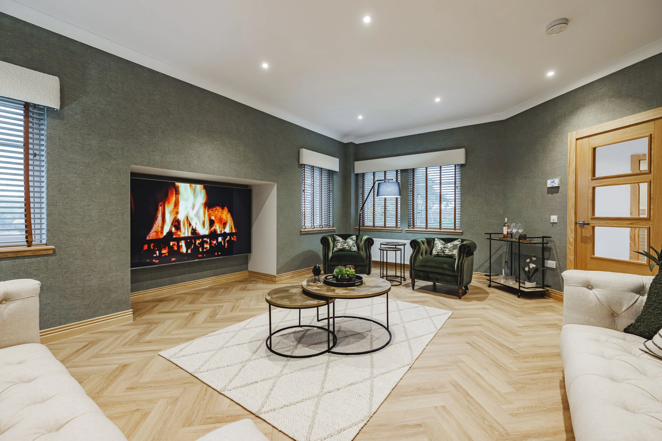 Living room with dark green walls, wood flooring, large windows with blinds, a fireplace, and white trim. Furniture includes green armchairs, white sofas, a round coffee table, and a side table with a lamp and drinks.
