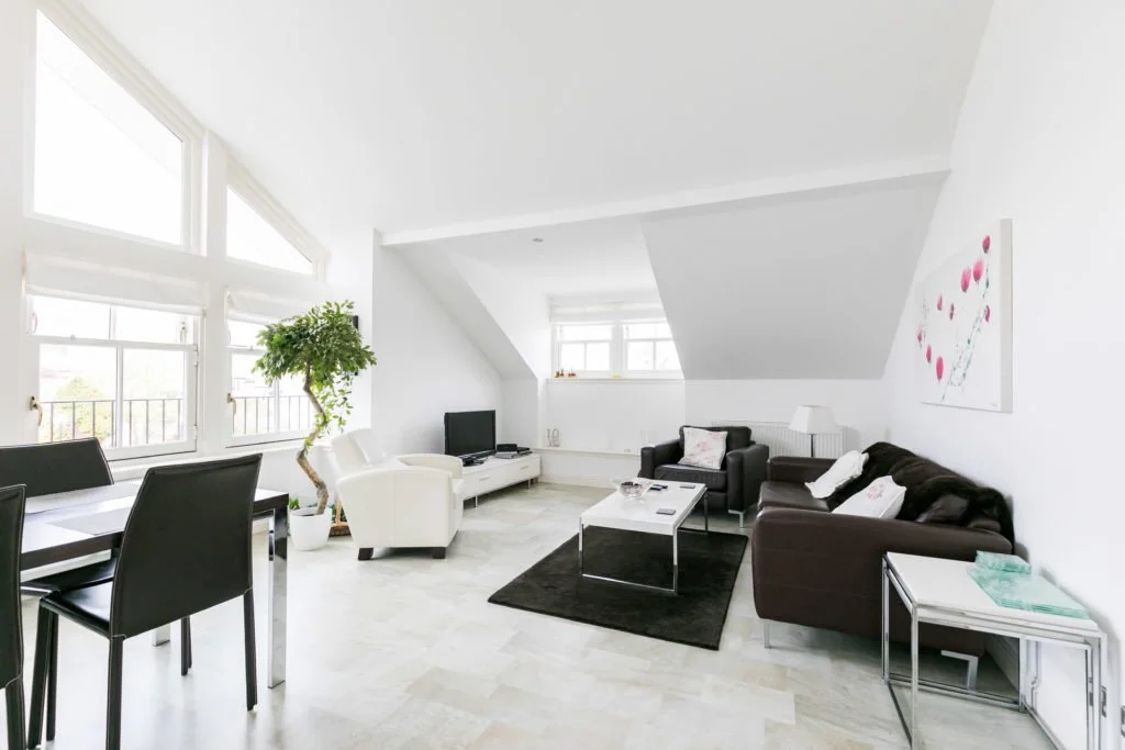 Bright living room with white walls and large windows, black and white sofas, a small black rug, a coffee table, a television, and a dining area with black chairs.