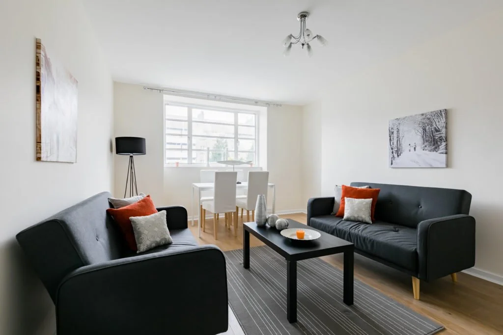 Living room with two dark gray sofas, a black coffee table, white walls, wooden floor, large window, dining table with four white chairs, black floor lamp, wall art, and decorative vases on the coffee table.