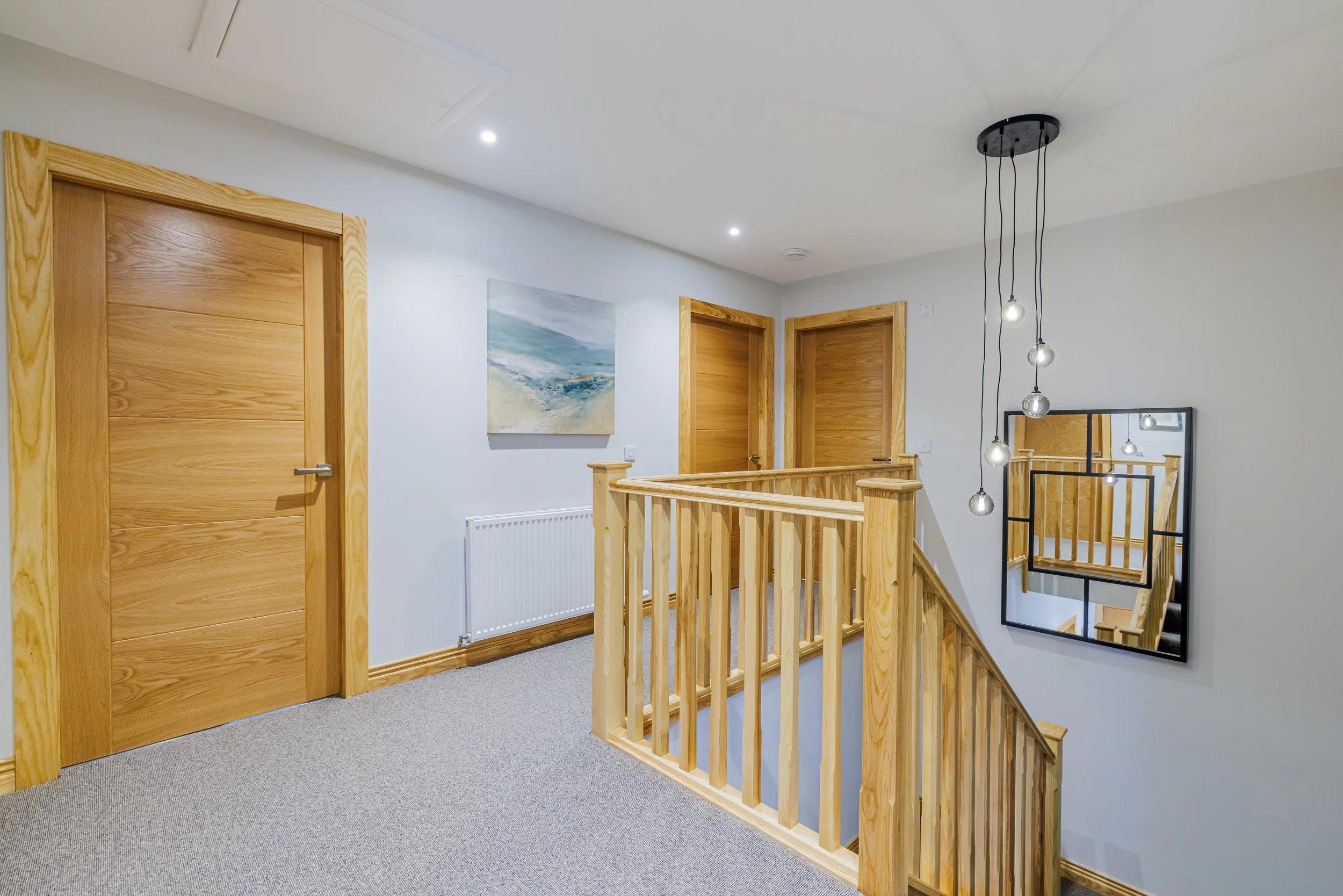 Interior view of a staircase landing with wooden doors, a painting, a mirror, and modern hanging lights.
