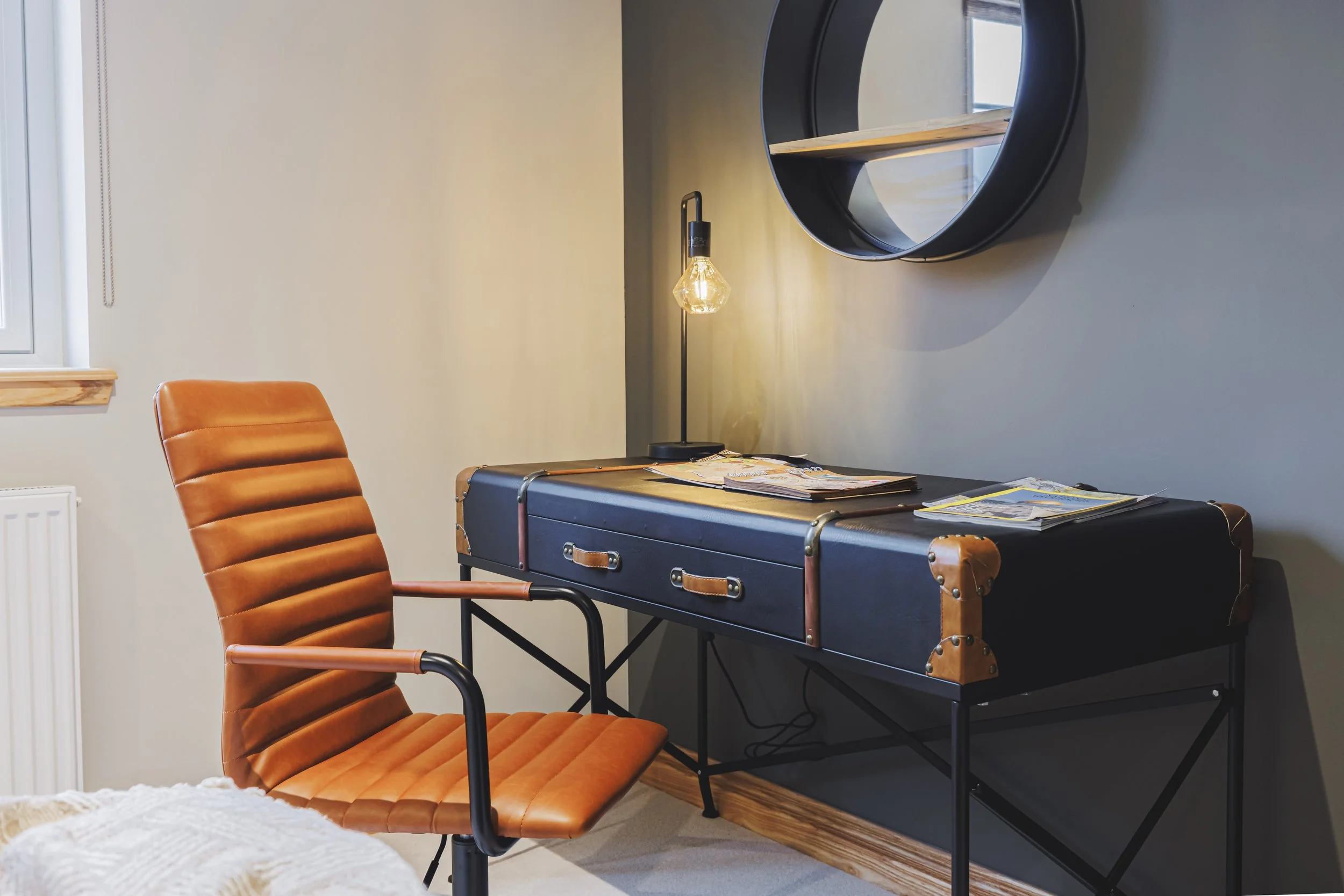 An office corner with an orange leather chair, a black desk with magazines, a modern desk lamp with an exposed light bulb, a round black mirror on a gray wall, a window with a wooden trim, and a beige wall.