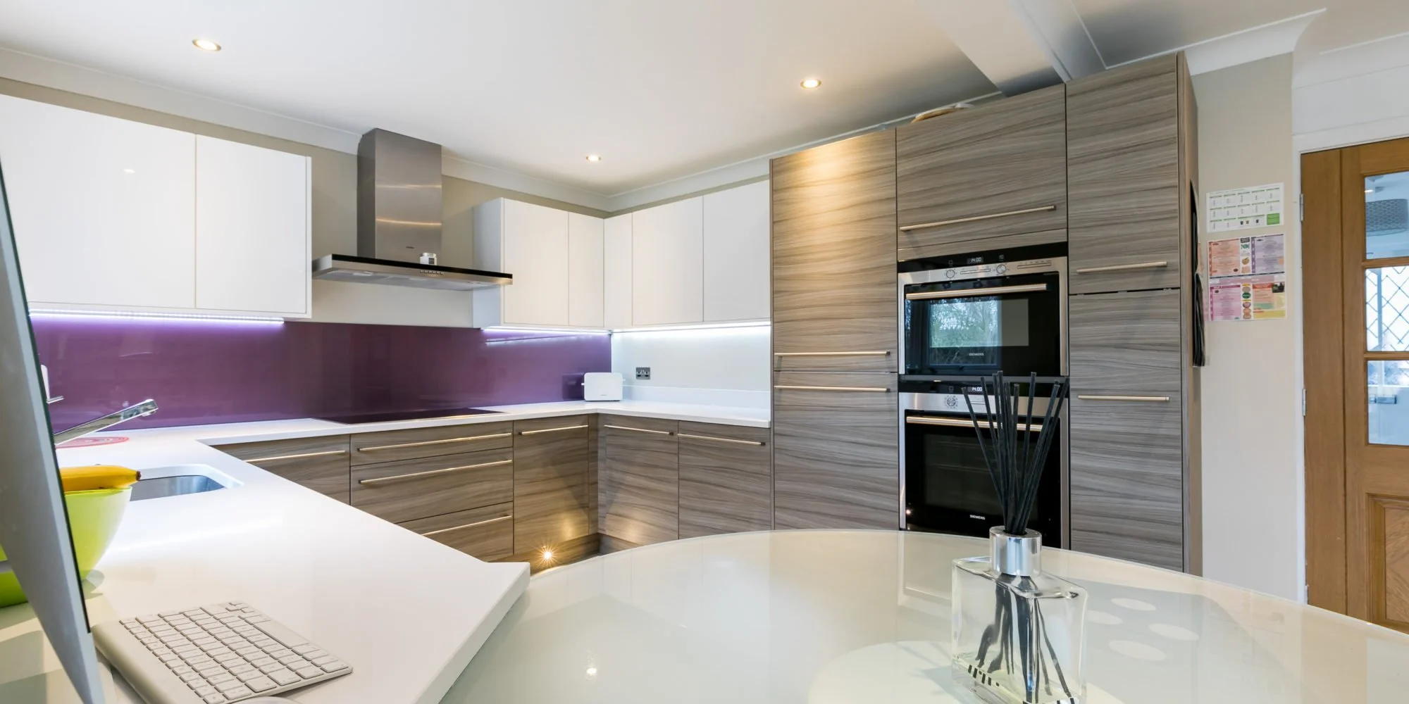 Modern kitchen with white and wood cabinetry, purple backsplash, stainless steel appliances, and a white countertop with a computer and yellow bowl.