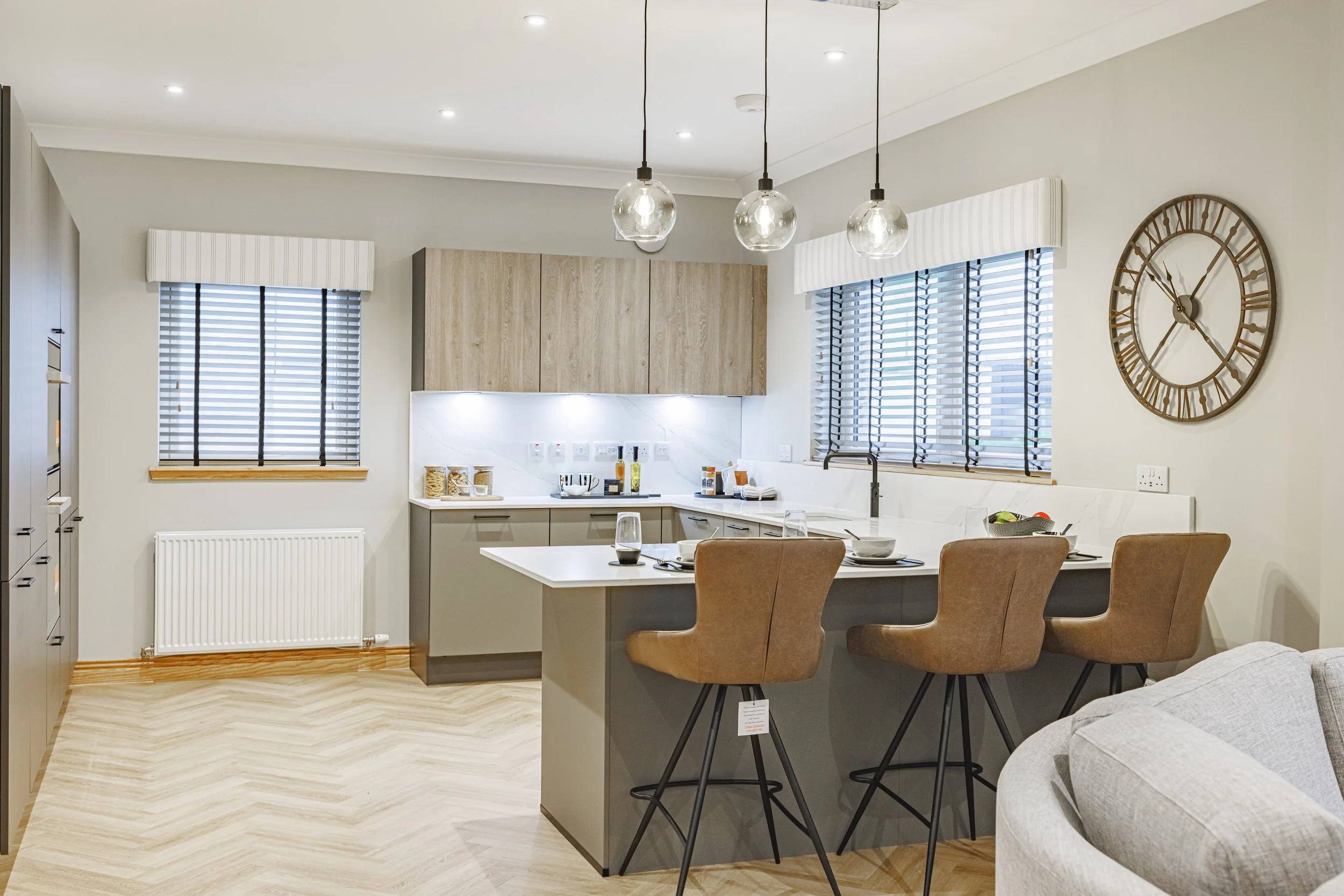 Modern kitchen and dining area with wooden and grey cabinetry, large windows with blinds, pendant lights, and wall clock.
