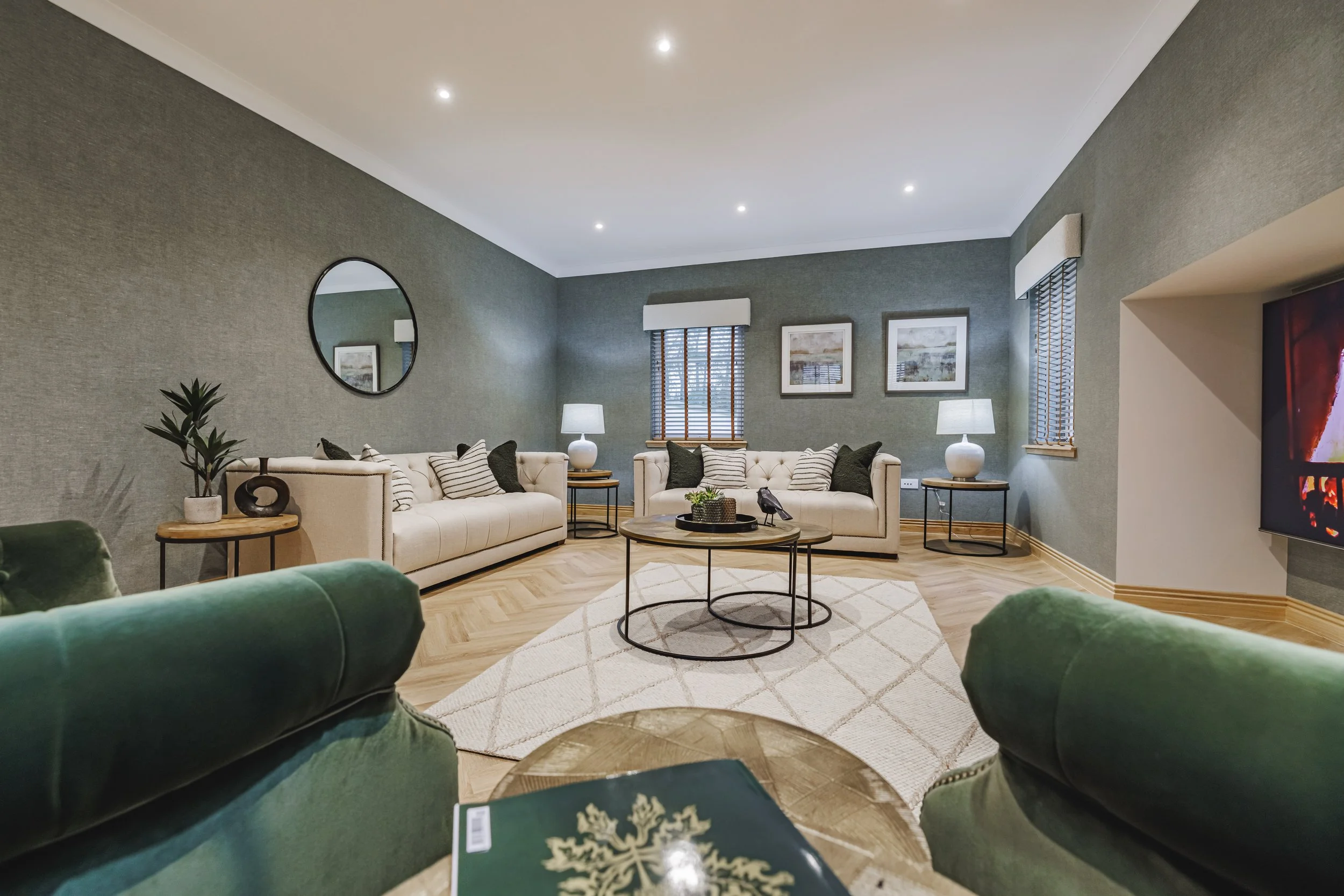 Living room with green velvet sofa, beige sofas with striped and solid cushions, round coffee table, side tables with lamps, decorative plant, wall art, window with blinds, mirror, and a fireplace.
