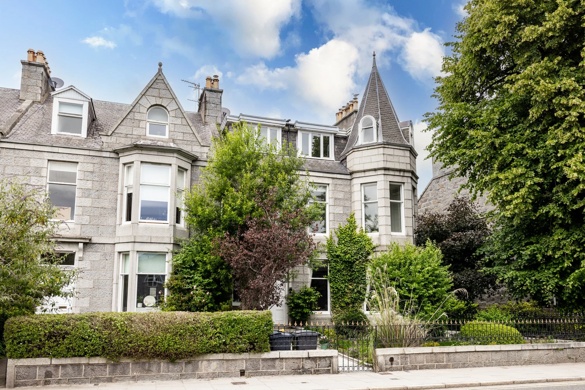 A large historical building with a stone facade, multiple windows, and steeply pitched roofs, surrounded by lush green trees and bushes under a partly cloudy sky.