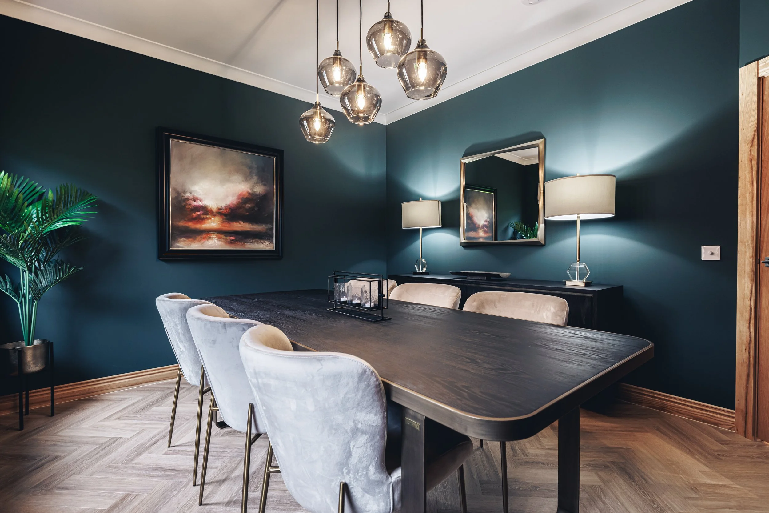 A modern dining room with a dark wooden table, six beige upholstered chairs, and decorative lighting. The walls are teal with a framed abstract painting and a large mirror. There are two table lamps and a plant in a pot.