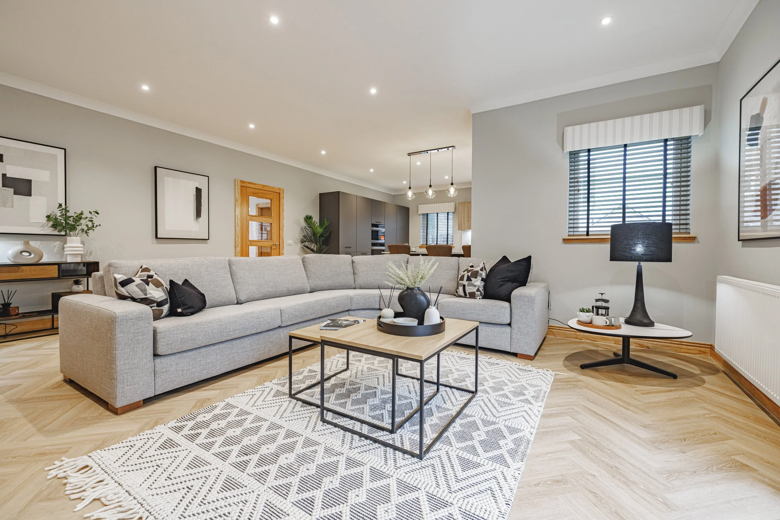 Living room with a light gray sectional sofa, black and white pillows, a wooden coffee table with vases and books, a patterned rug, artwork on the walls, a black lamp, and a window with blinds. The background shows a kitchen with gray cabinets and pendant lighting.