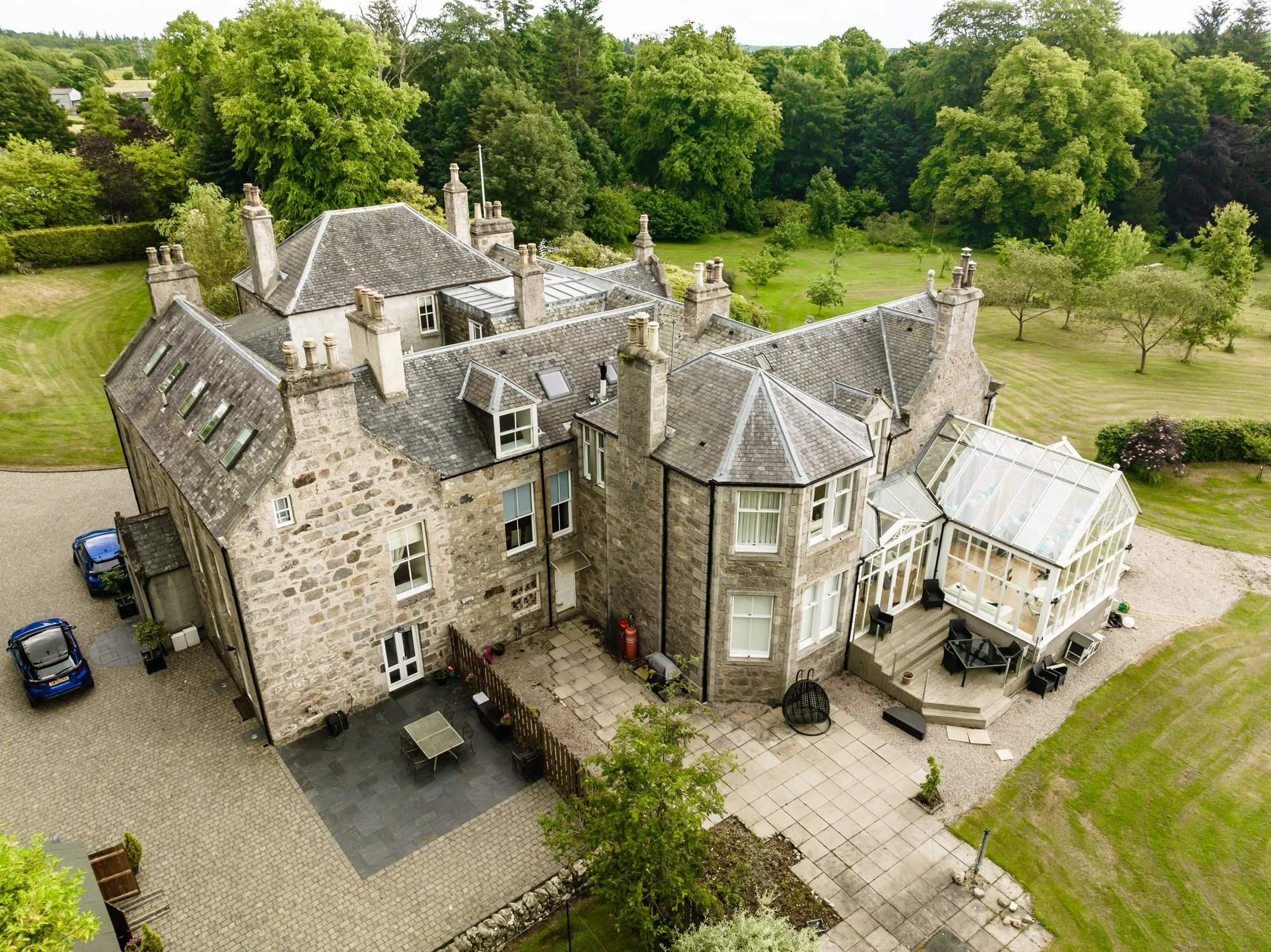 A large stone mansion with multiple chimneys and gray shingle roofs seen from above, surrounded by green trees and a neatly manicured lawn, with a paved patio area and outdoor furniture.