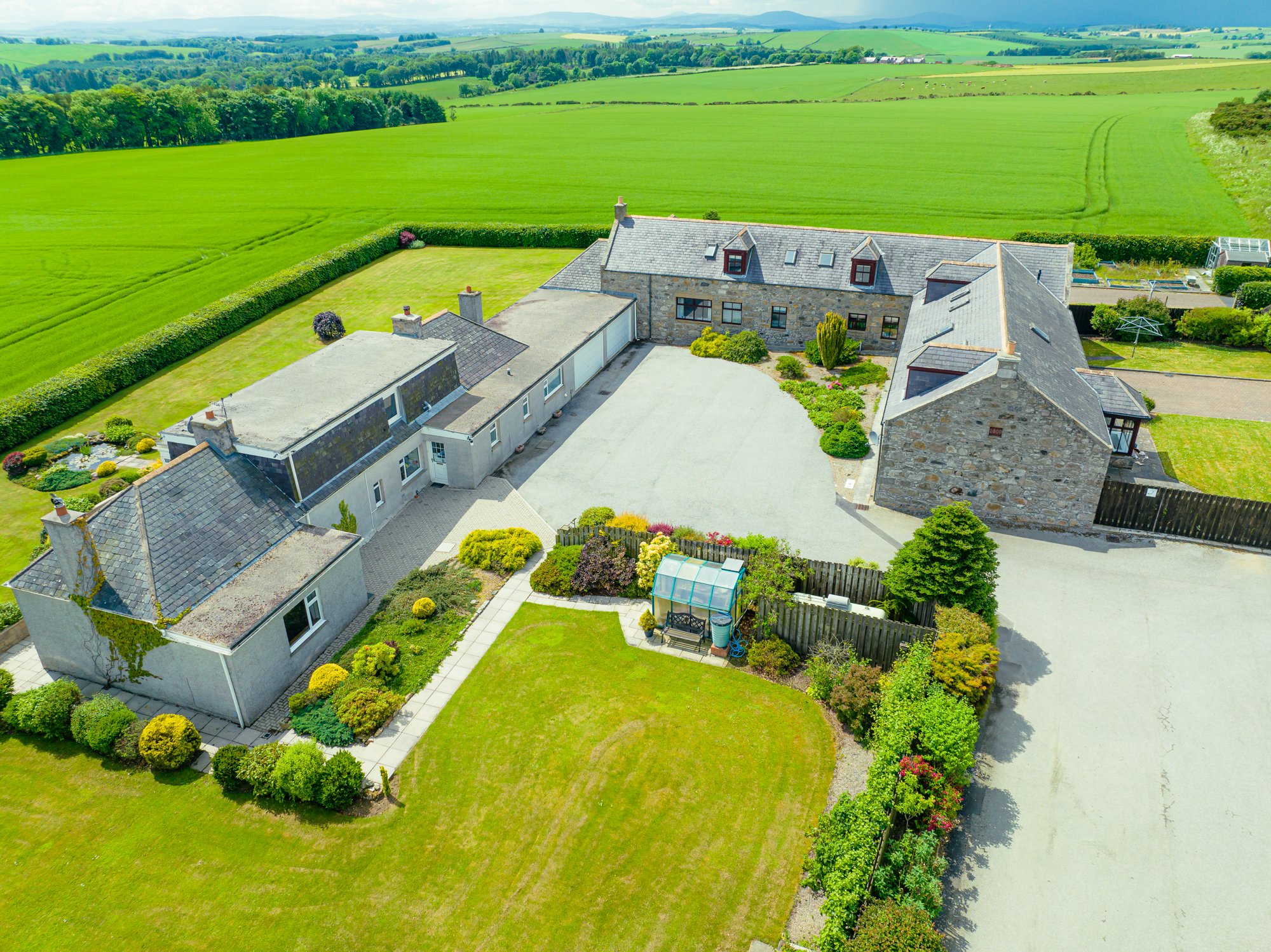 Aerial view of a stone house with a gray roof, surrounded by a lush green lawn and garden, with expansive farmland and fields in the background.
