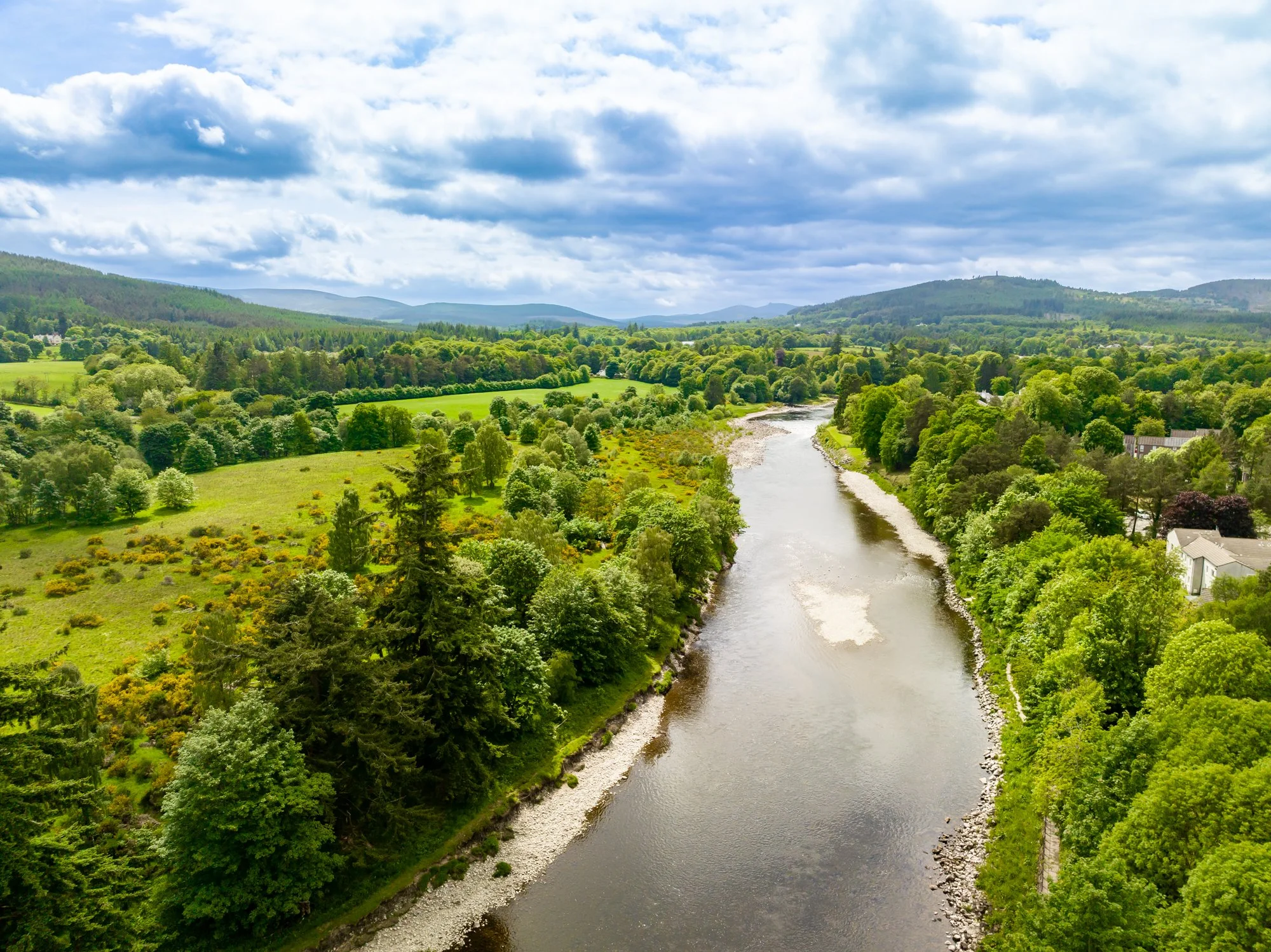 A scenic river flowing through lush green forested landscape with rolling hills and partly cloudy sky.