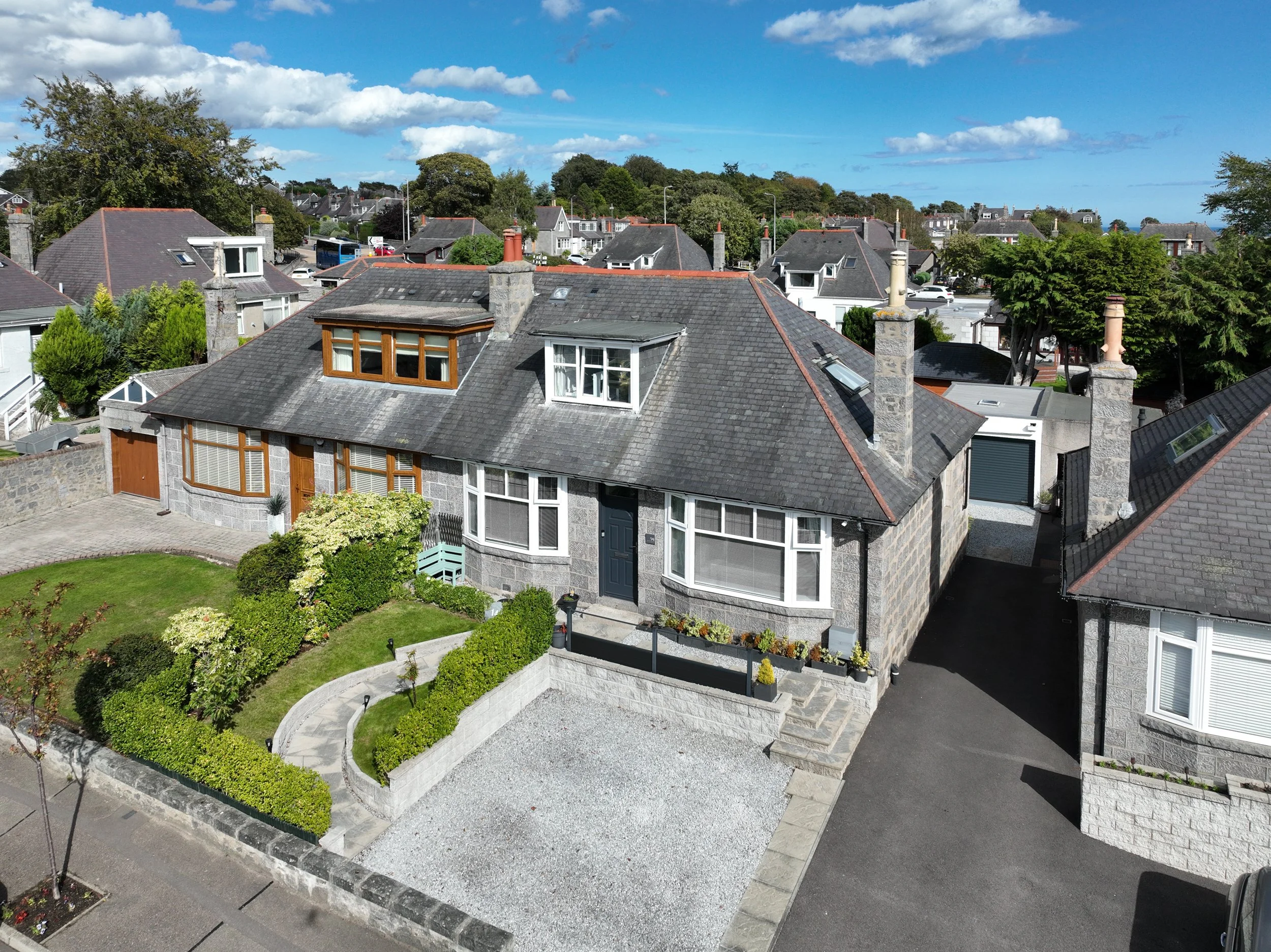 Aerial view of a residential neighborhood with multiple houses featuring gray roofs, chimneys, dormer windows, and well-maintained gardens under a partly cloudy sky.