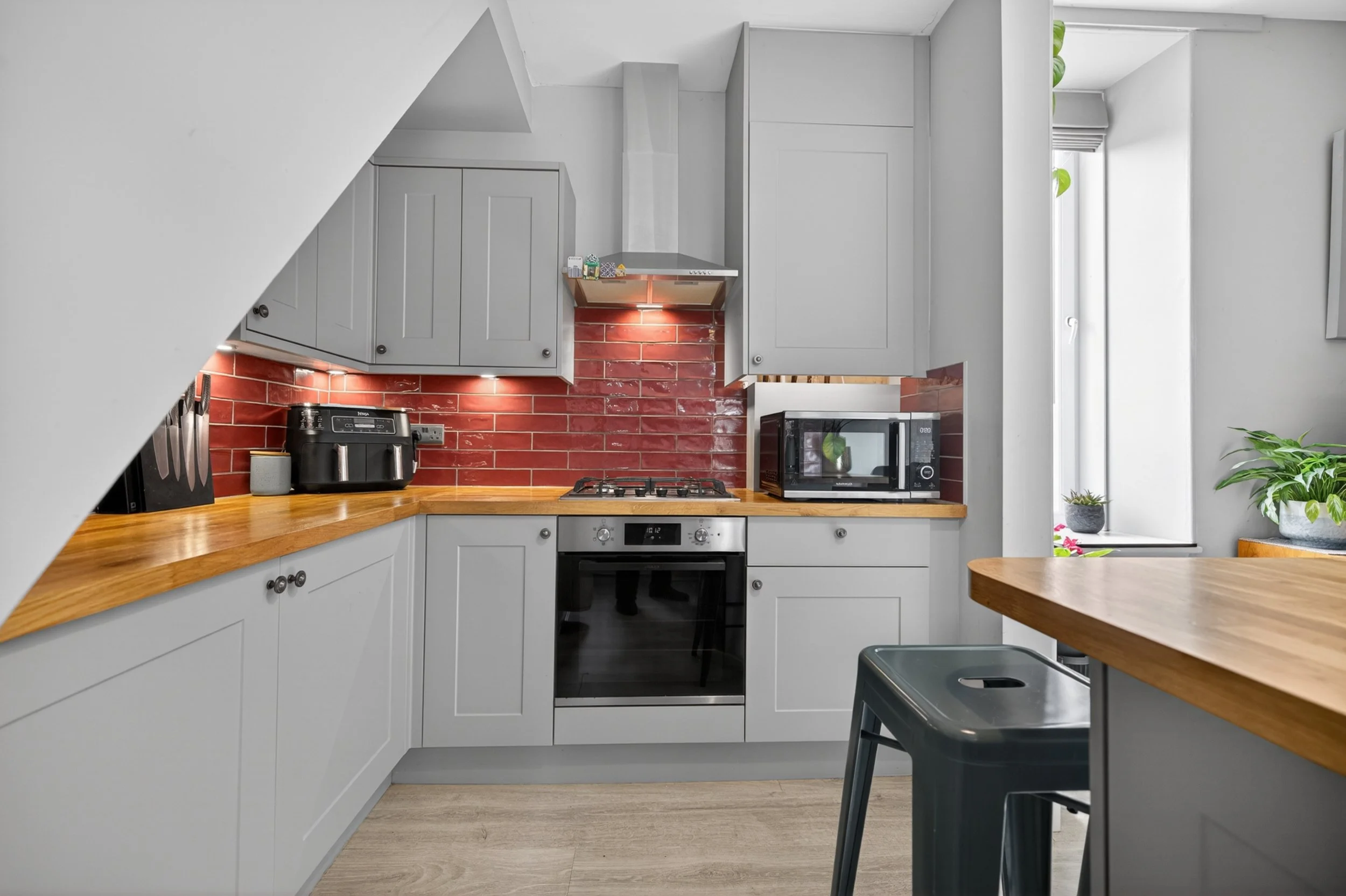 Modern kitchen with gray cabinets, a red brick backsplash, and wooden countertops. Appliances include a microwave, oven, and coffee maker. There is a small table with two black chairs, and a window with plants nearby.
