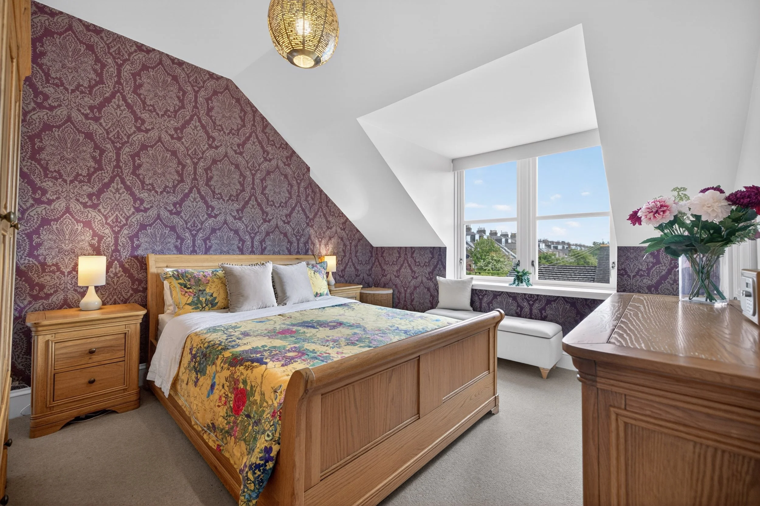 A cozy bedroom featuring a wooden bed frame with floral and neutral bedding, a purple damask accent wall, a round ceiling light, large windows with a view of rooftops and blue sky, a wooden nightstand with a lamp, and a bouquet of pink and purple flo