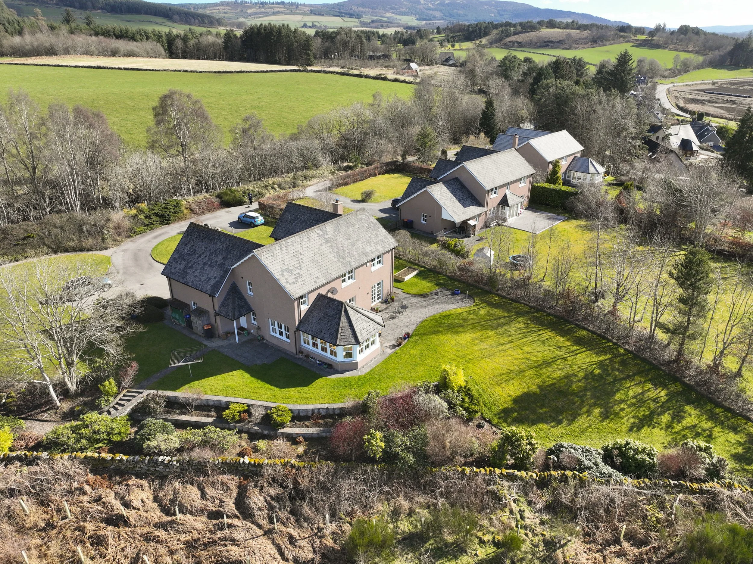 Aerial view of a residential neighborhood with houses, green lawns, and trees amid hilly landscape and farmlands.