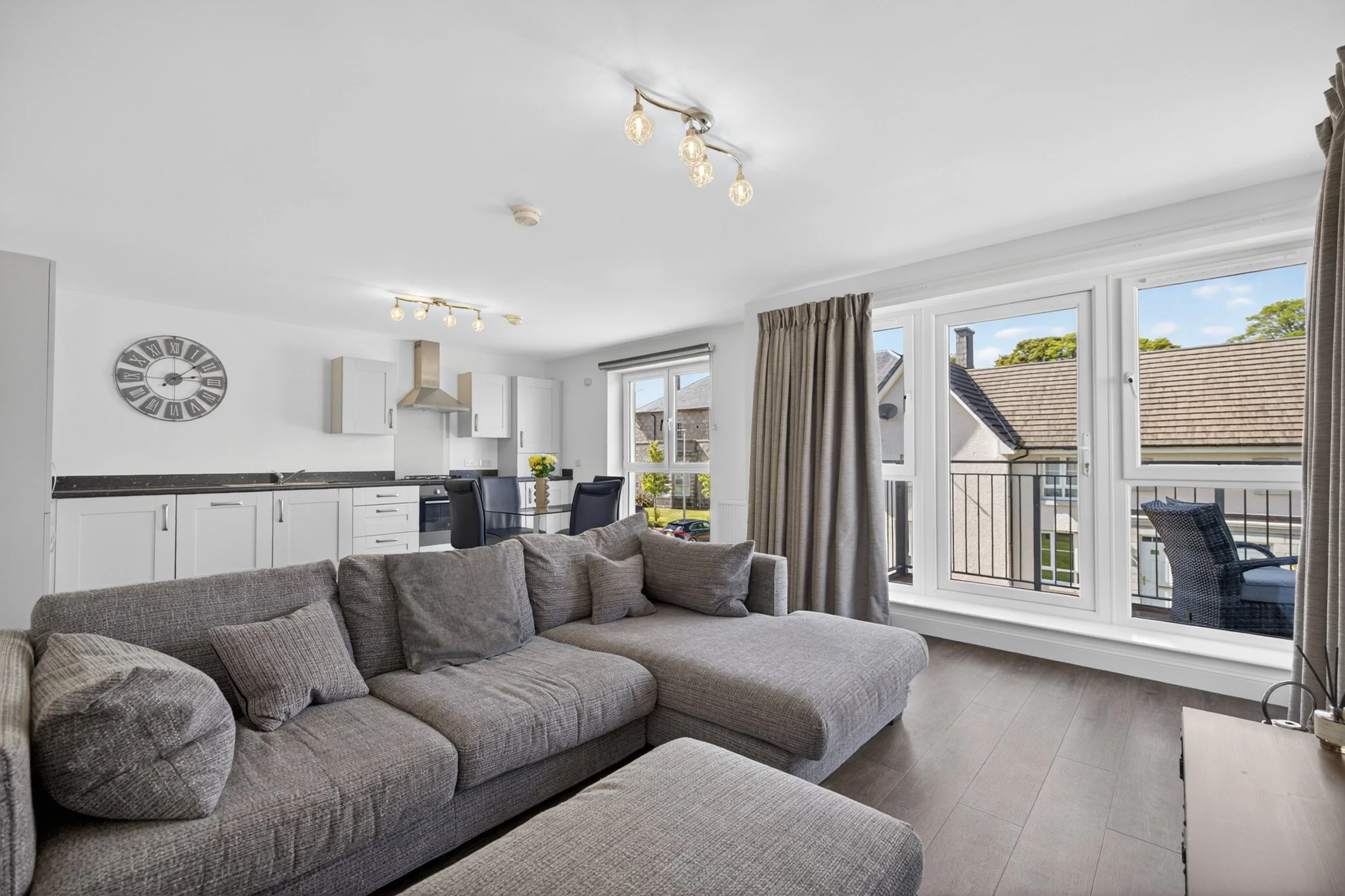 Living room with large windows, gray sofa, checkered pillows, kitchen with white cabinets, wall clock, ceiling lights, and outdoor view of rooftops and trees.