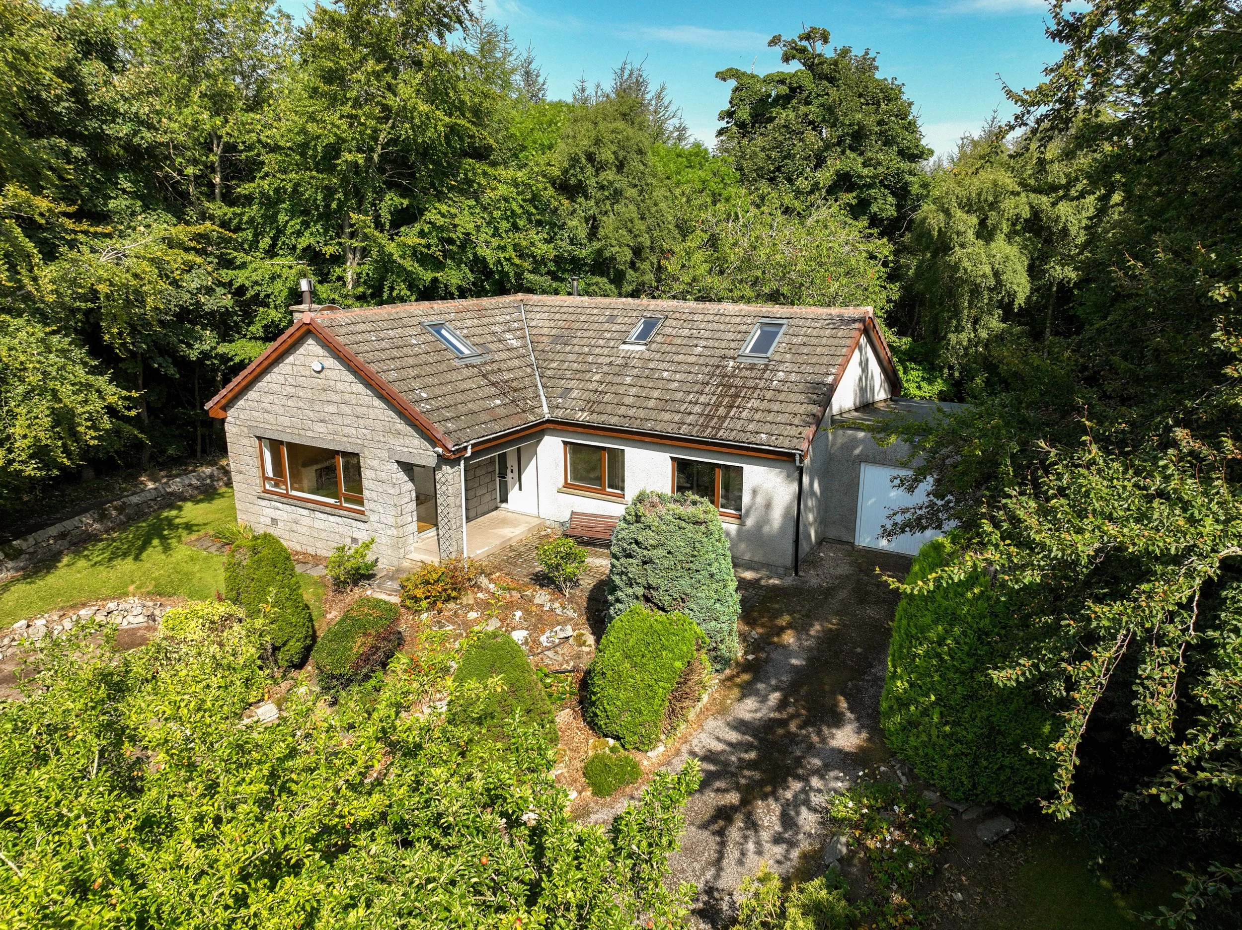 A house with a gable roof and three skylights, surrounded by trees and a small garden with bushes and a lawn, under a bright blue sky.
