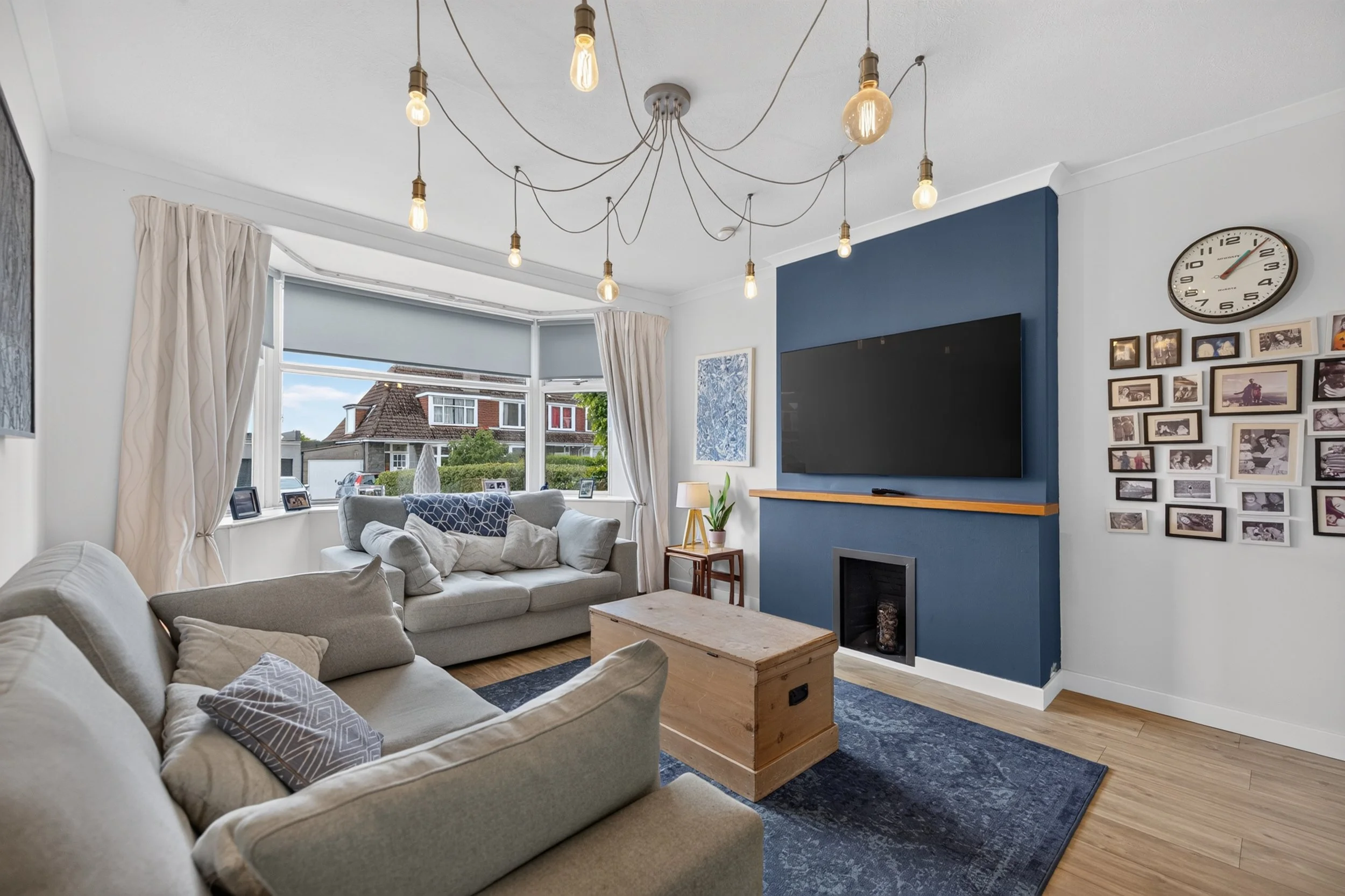 Living room with beige sofas, a wooden coffee table, a large window with curtains, a wall-mounted TV, a gallery wall with framed photos, a clock, and a modern chandelier with exposed light bulbs.