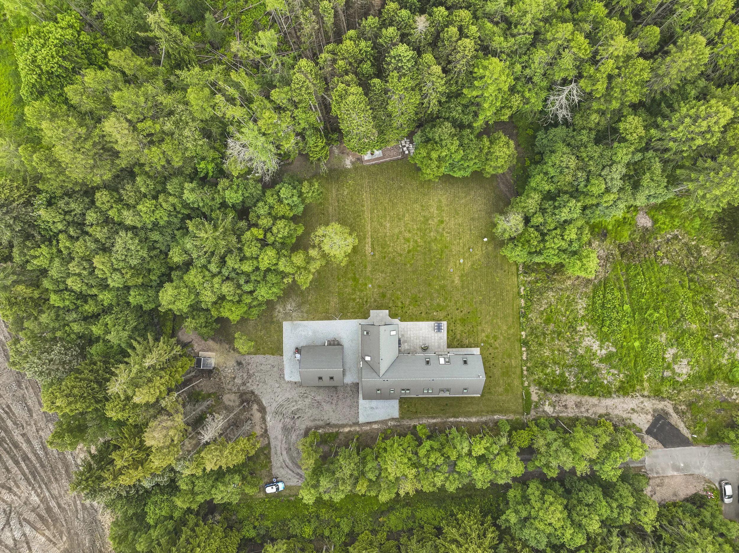 An aerial view of a house surrounded by dense green trees and a large open grassy yard.