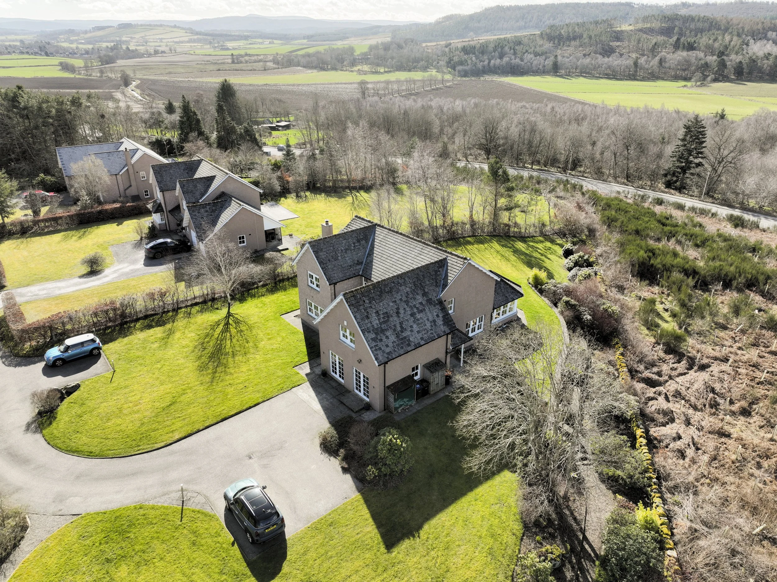 Aerial view of a residential neighborhood with modern beige houses, green lawns, and parked cars, surrounded by leafless trees and open farmland in the distance.