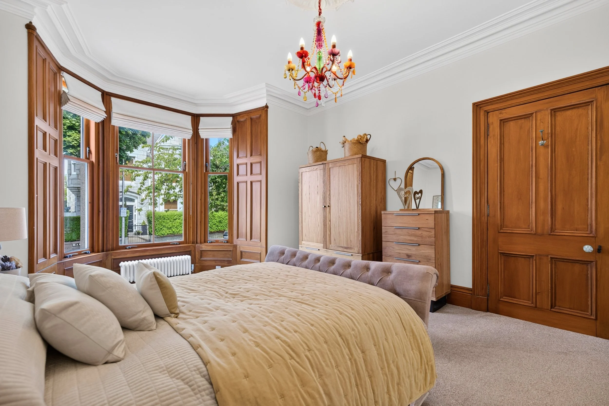 A bedroom with large bay windows featuring white blinds and wooden trim, a beige tufted bed with cream pillows and a beige quilt, a wooden wardrobe, a dresser with decorative signs, a mirror, and a colorful chandelier hanging from the ceiling.