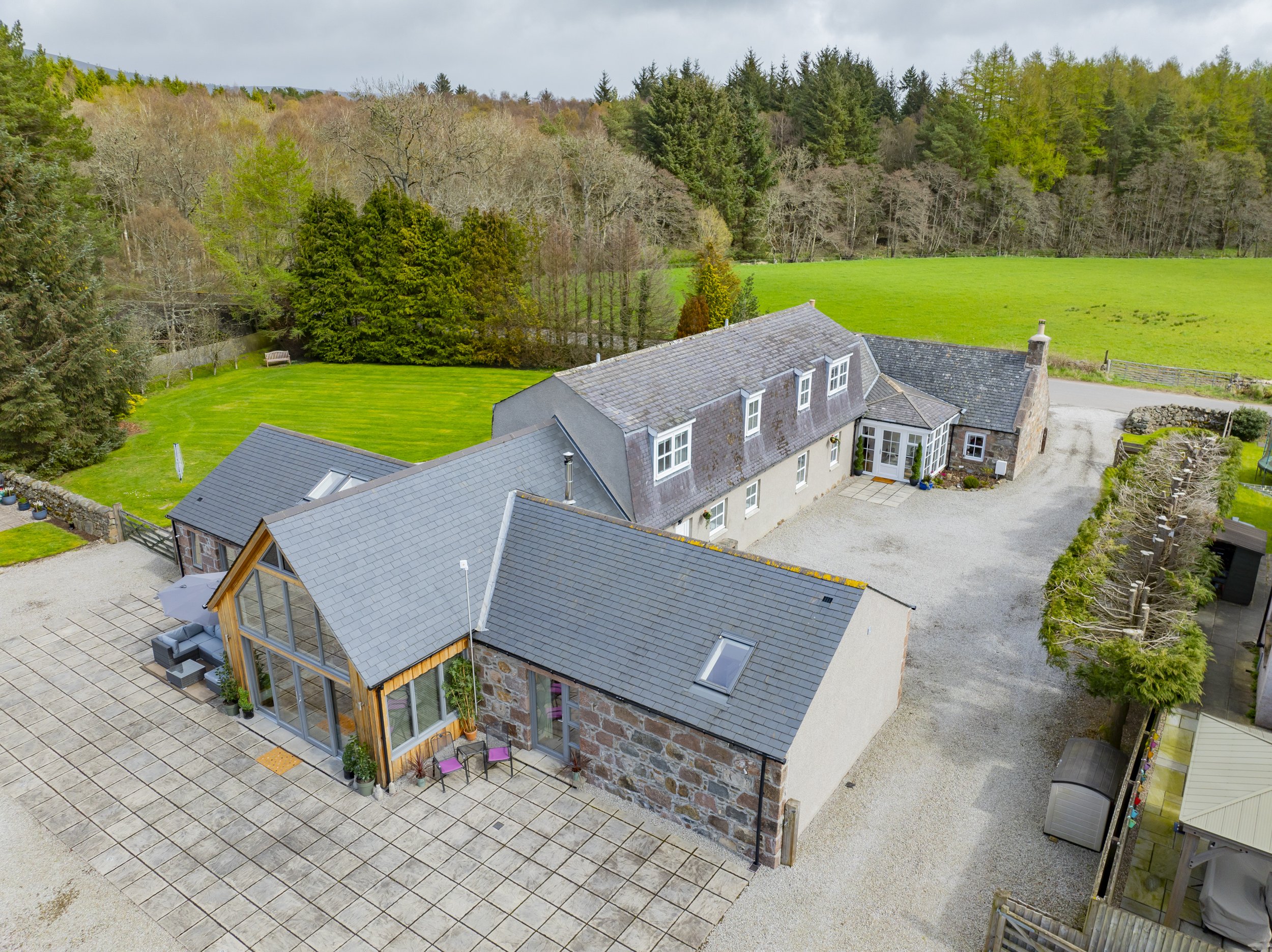 Aerial view of a stone house with a gray shingled roof, large glass-enclosed porch, and outdoor patio area. The house is surrounded by a green lawn, trees, and a gravel driveway leading to the property. In the background, there is a forest with decid