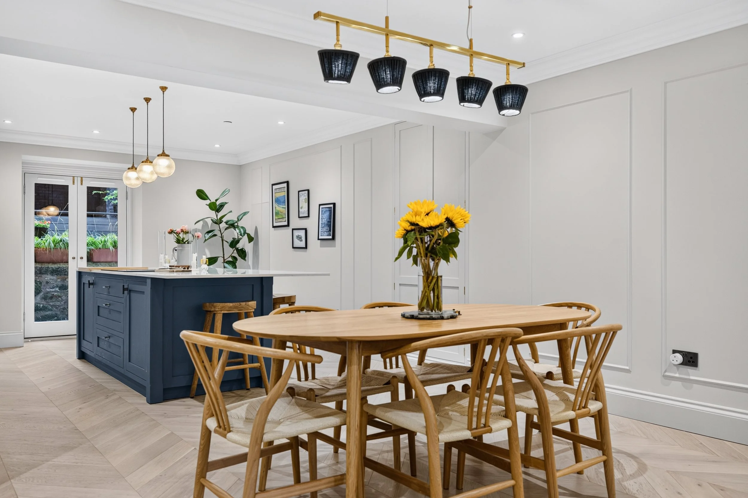 A dining area with a wooden table surrounded by wooden chairs, a vase with yellow sunflowers on the table, and a kitchen island with a blue base and white countertop. There are pendant lights hanging from the ceiling, artwork on the wall, and French 