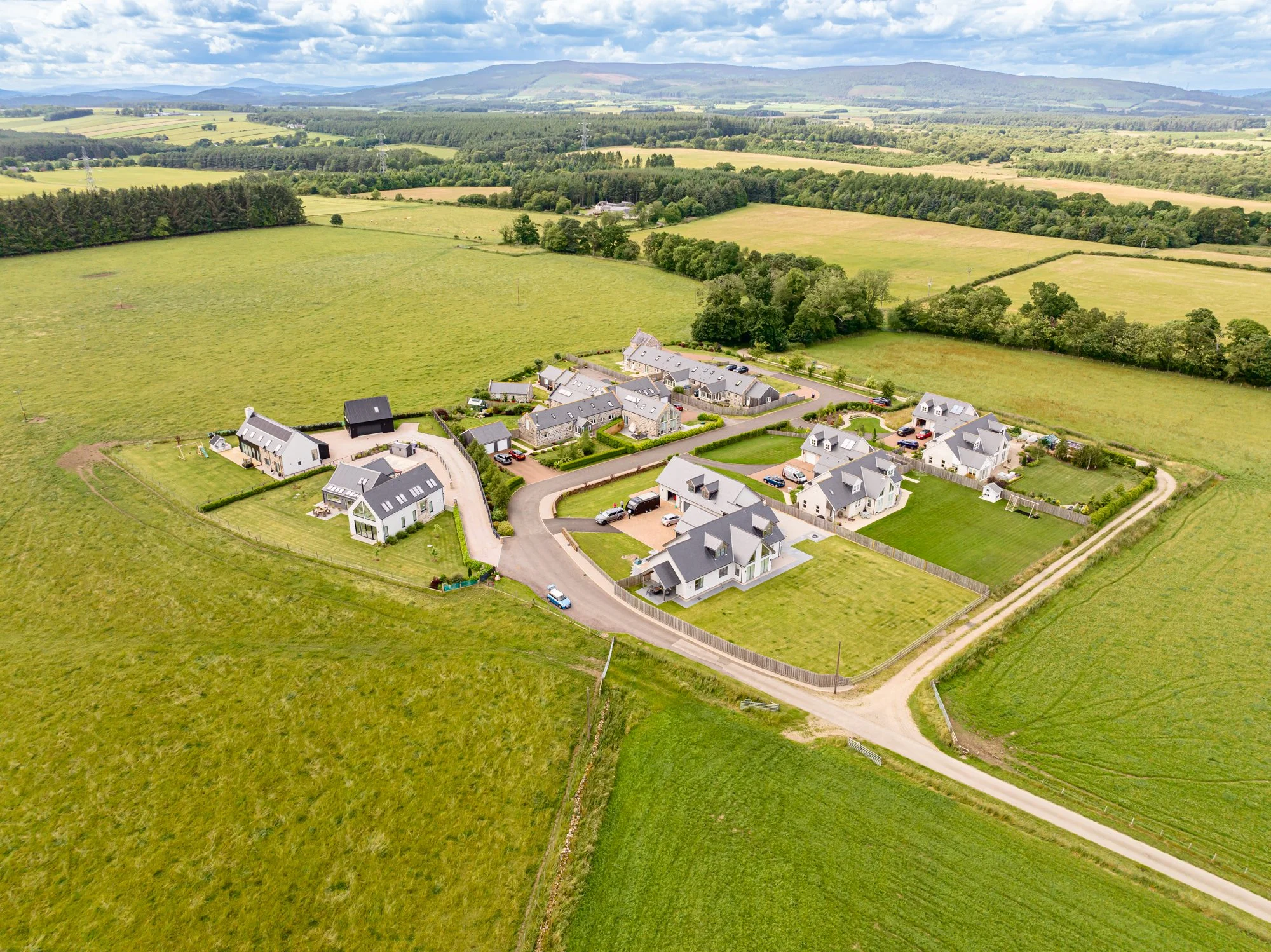 Aerial view of a new housing development with modern white houses, surrounded by green fields and farmland, with hills in the background under a partly cloudy sky.