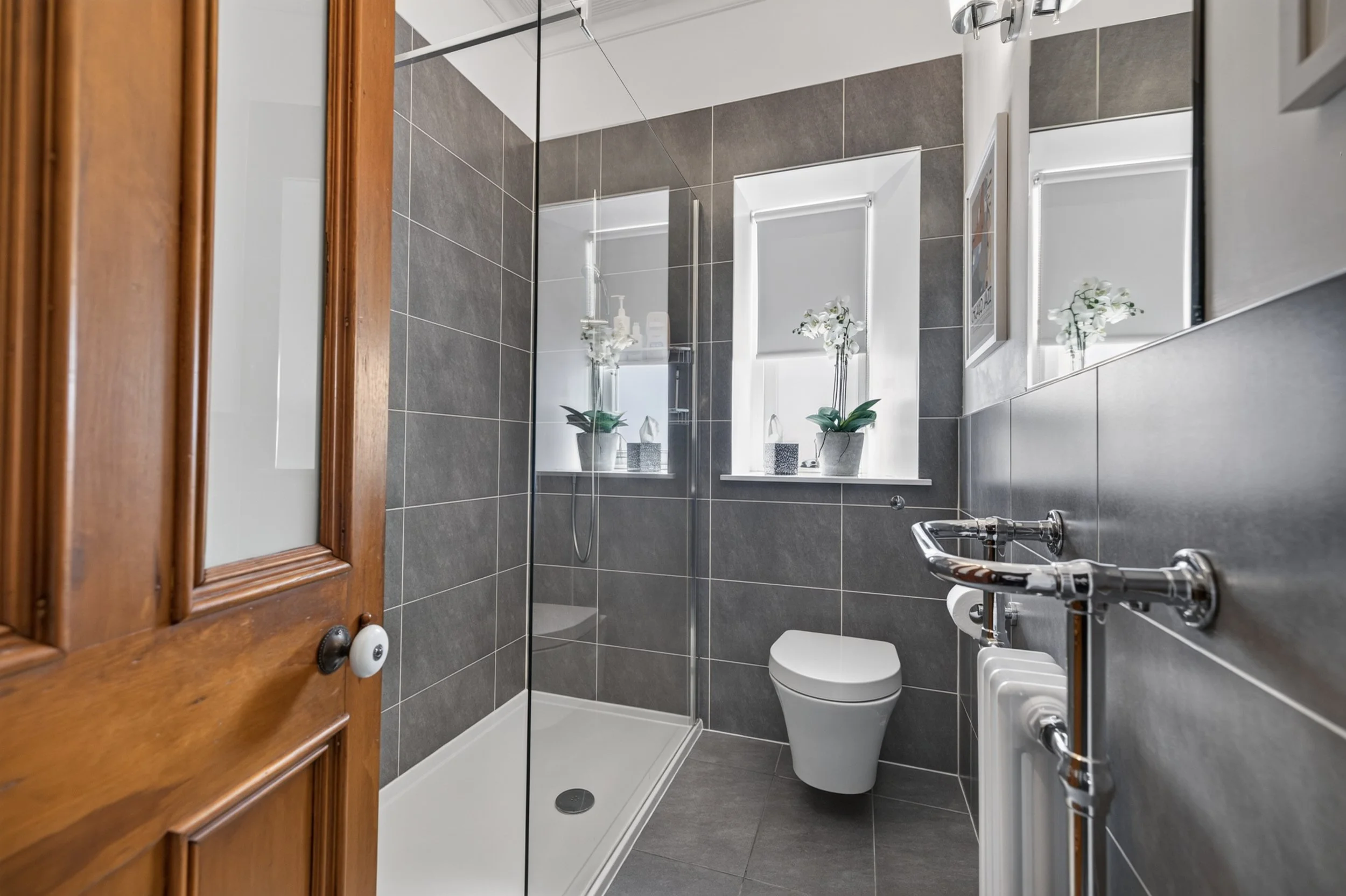 A modern bathroom with gray tiled walls, a white toilet, a shower area with glass door, and two windows with white blinds. There are white orchids in pots on the windowsill, and a wooden cabinet door is visible on the left.