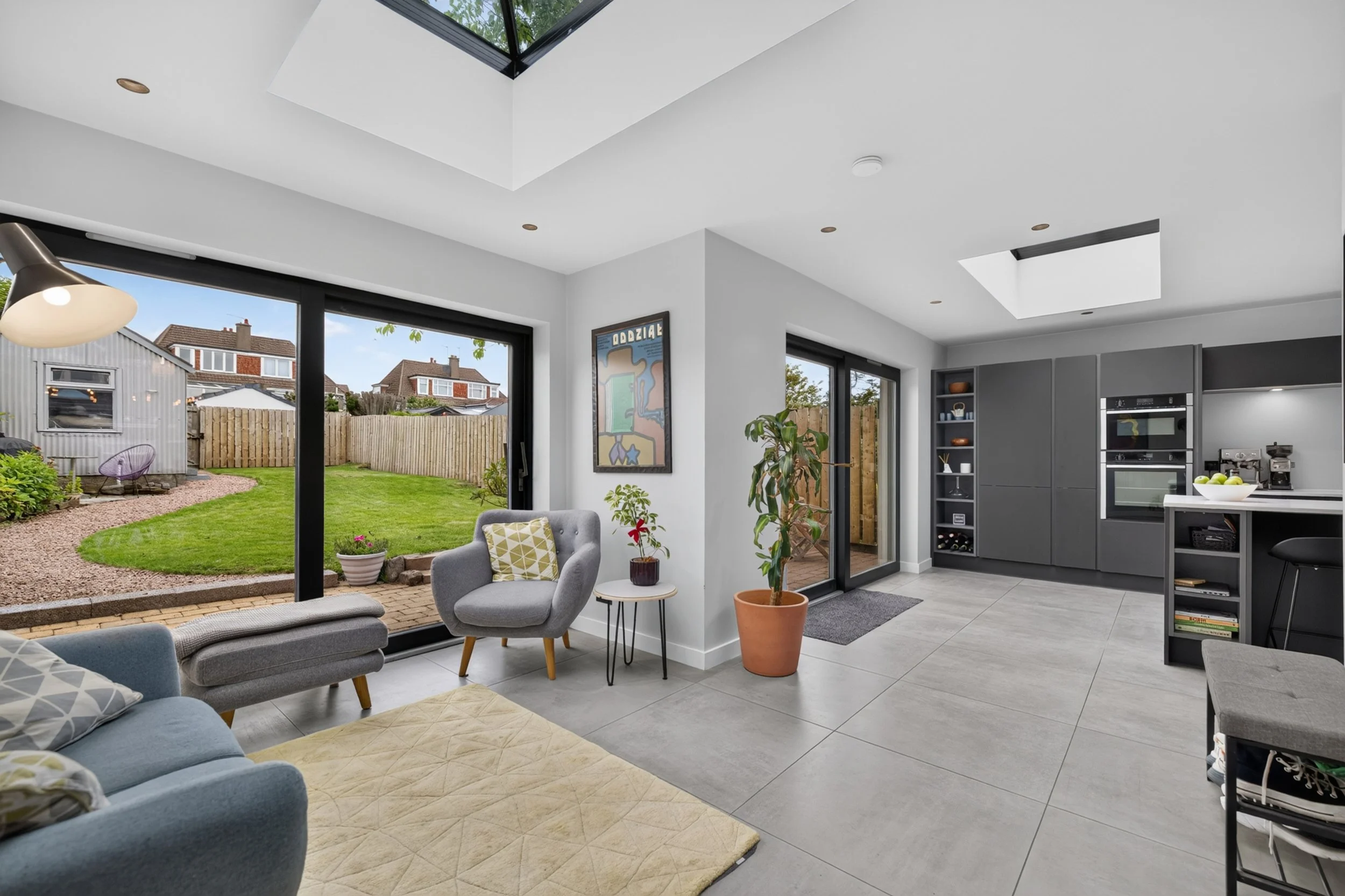 A modern indoor living room with large sliding glass doors opening to a backyard with green grass, a wooden fence, and neighboring houses. The room features a gray sofa, armchairs, potted plants, and artwork on the walls, with a kitchen visible in th