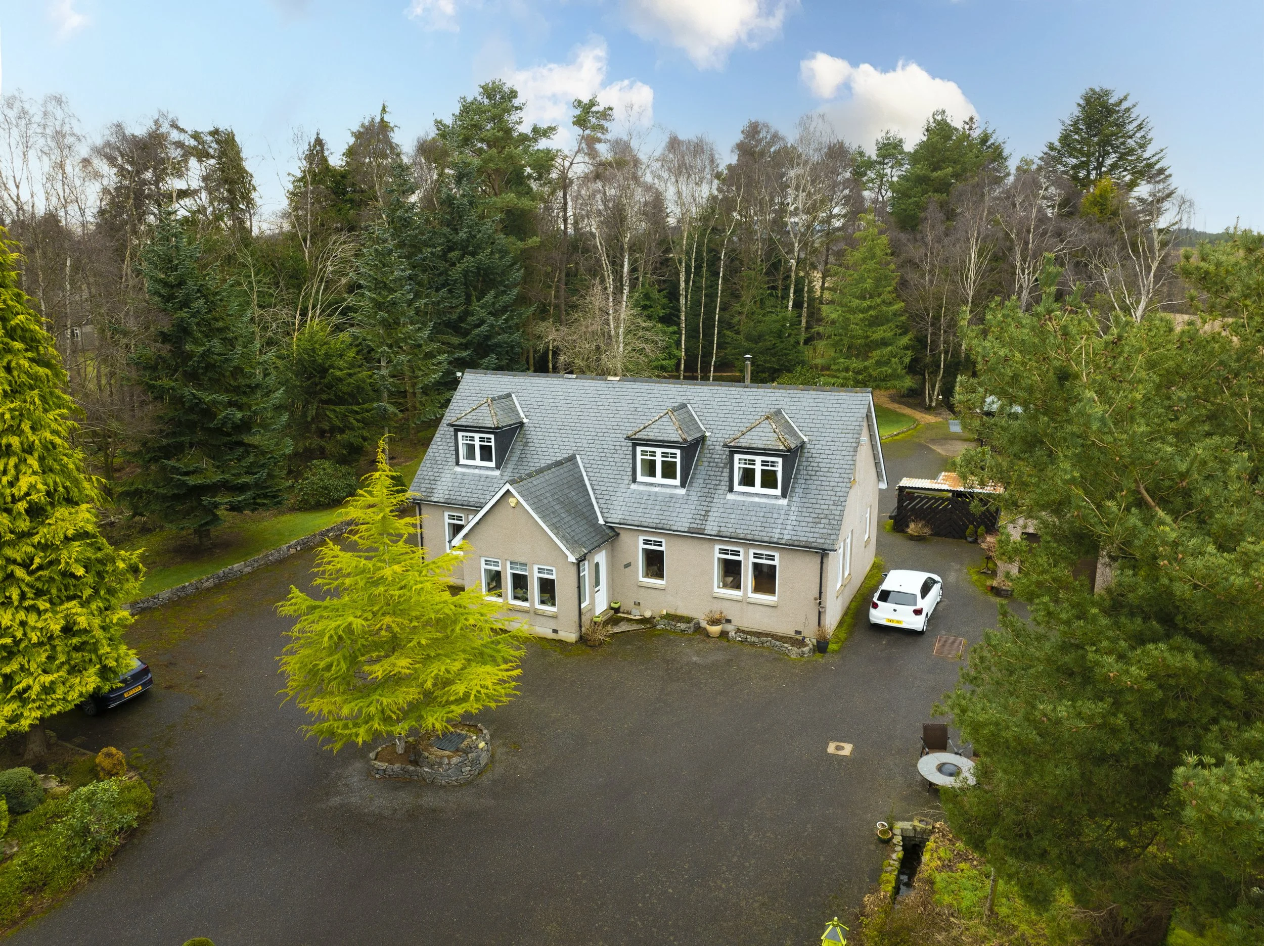 A house with a gray roof surrounded by many trees and a spacious yard, with several parked cars and a clear sky with clouds.