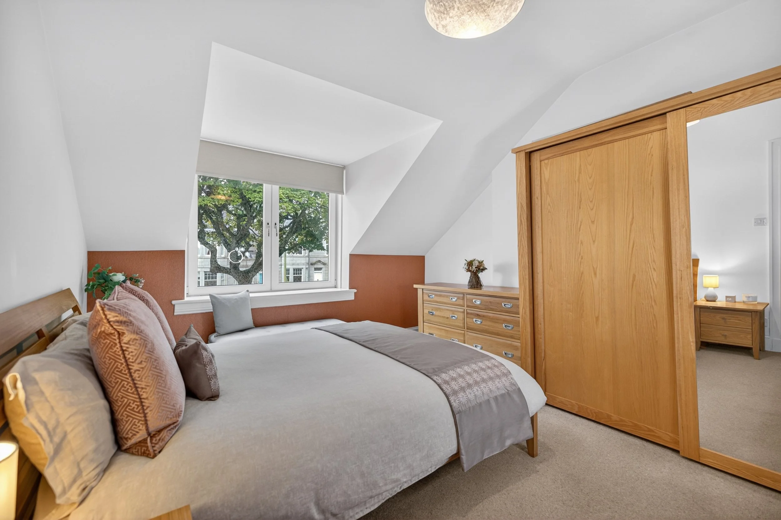 A modern bedroom with a large bed featuring multiple pillows, a window with a tree view, a wooden dresser, and a wooden sliding door.