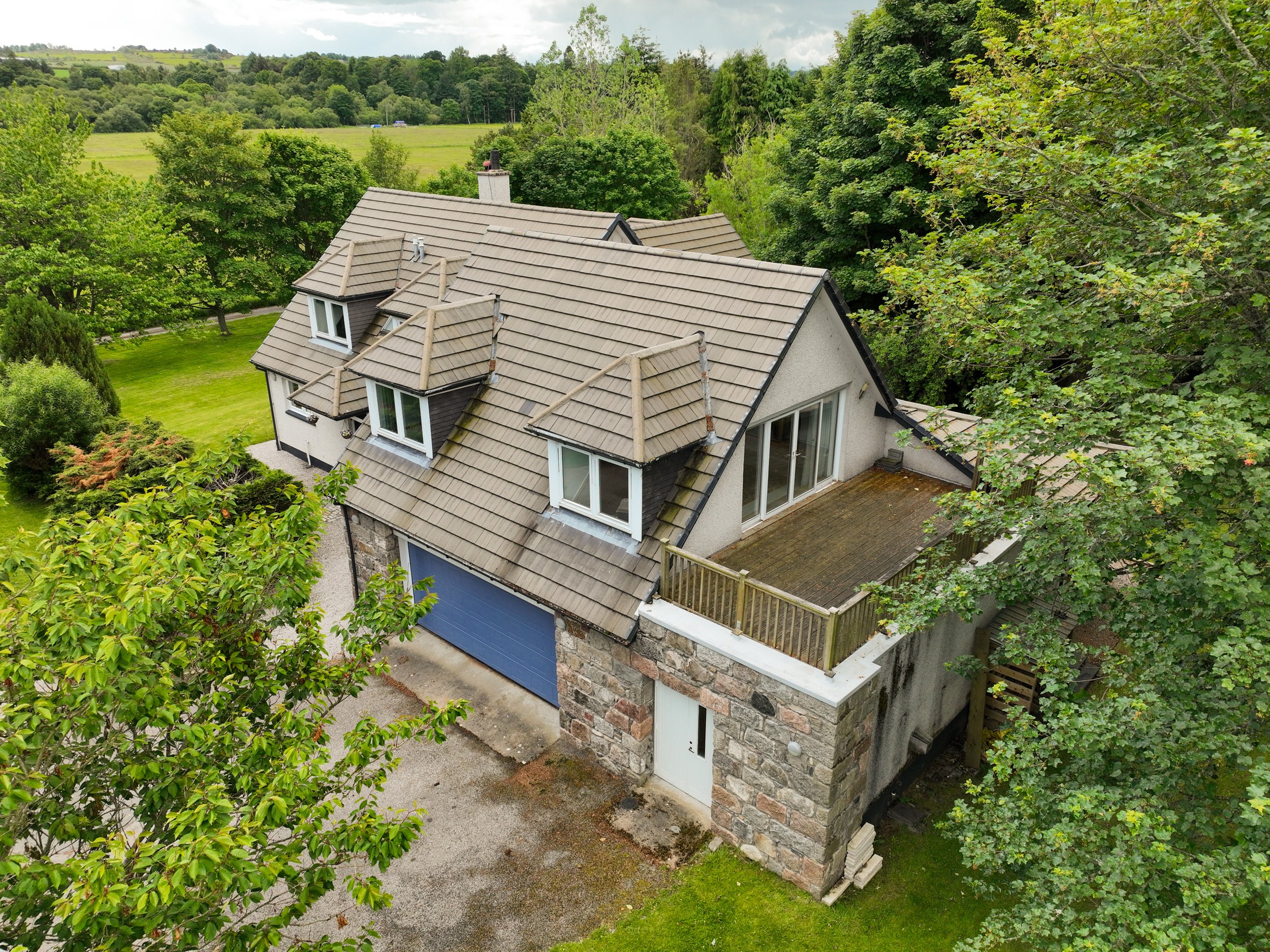 Aerial view of a house with a gray tile roof, multiple gables, white-framed windows, and a stone exterior wall, surrounded by lush green trees and a well-maintained yard with a blue garage door.